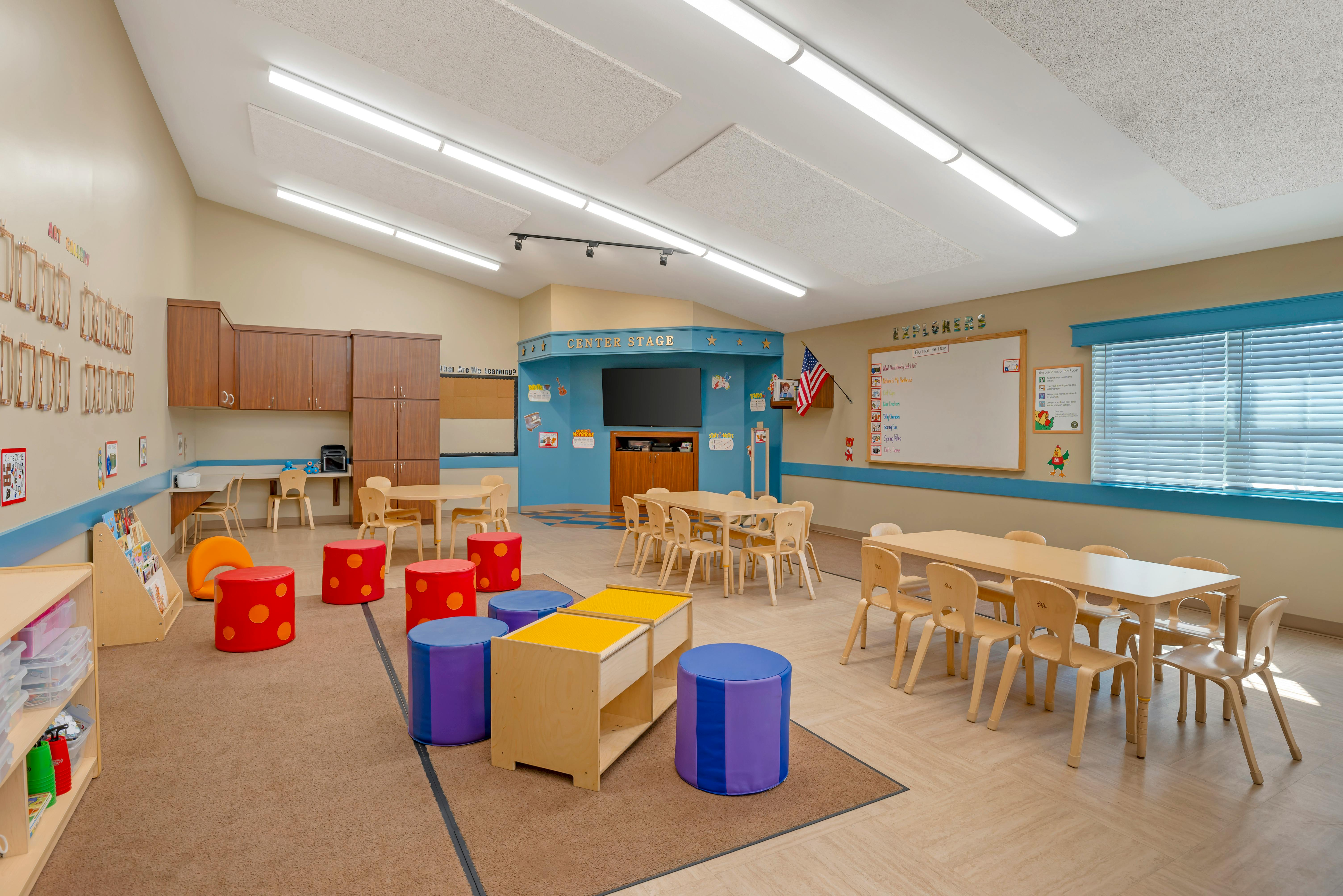 Classroom with small tables, chairs, and play items, with "CENTER STAGE" over a screen