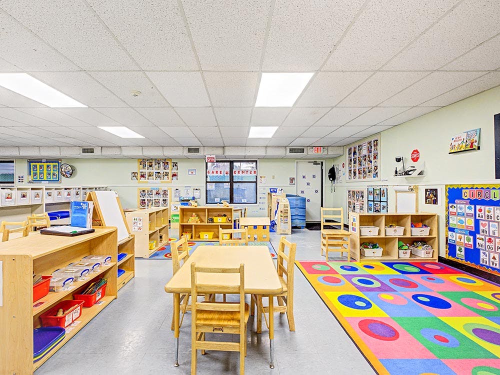 Empty classroom with children's tables and chairs, shelves, and a colorful rug