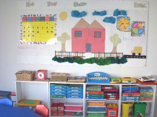 Classroom wall with a calendar, paper house, and shelves with learning materials