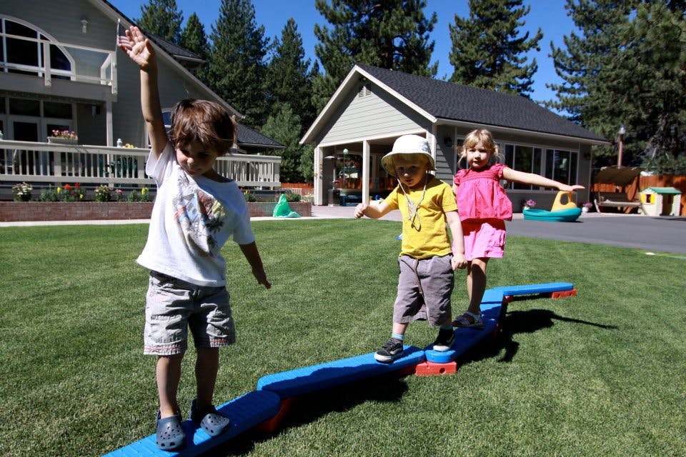 Three children playing on a balance beam on a grassy lawn