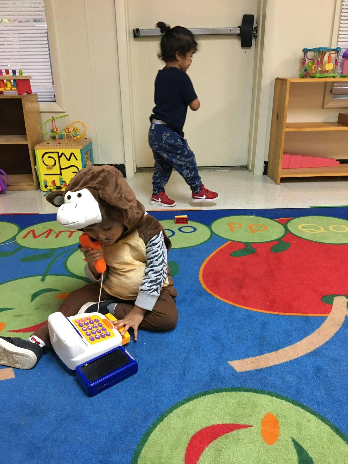 Child in monkey costume playing with toy phone, another child walking in background