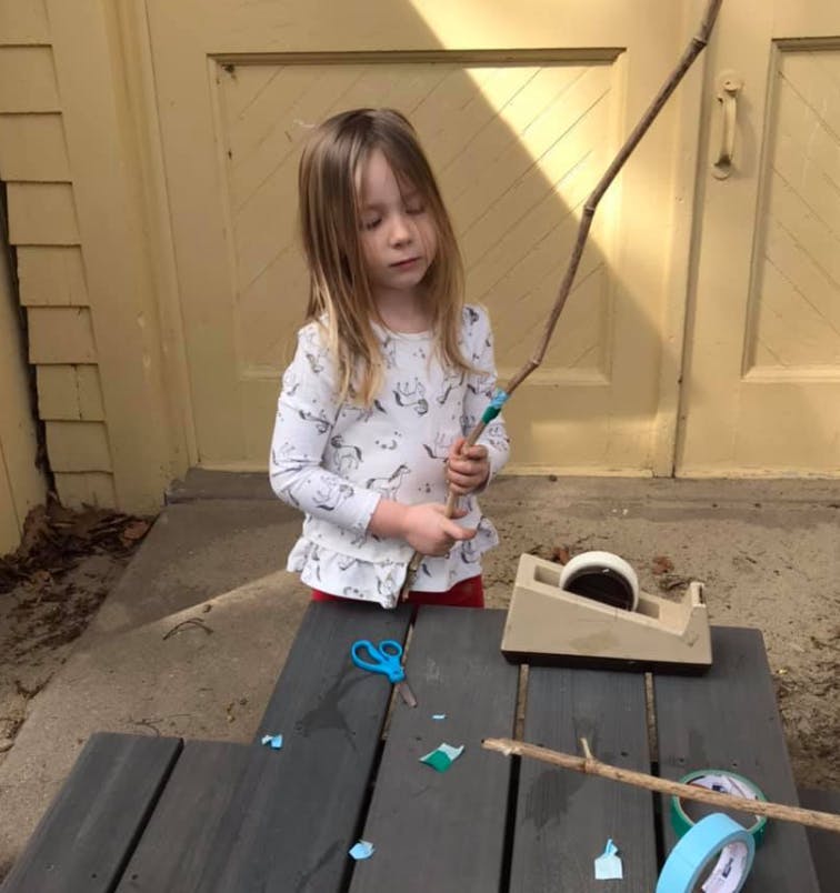 Child holding a stick with tape, with scissors and tape dispenser on a table