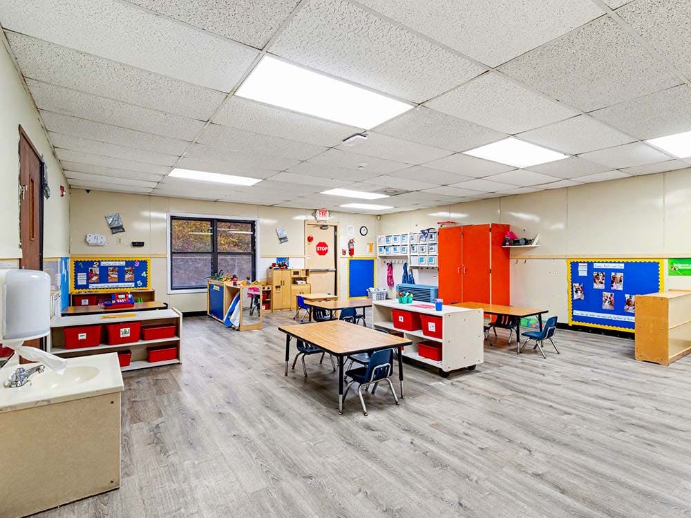 Empty preschool classroom with small tables and chairs, bulletin boards, and storage