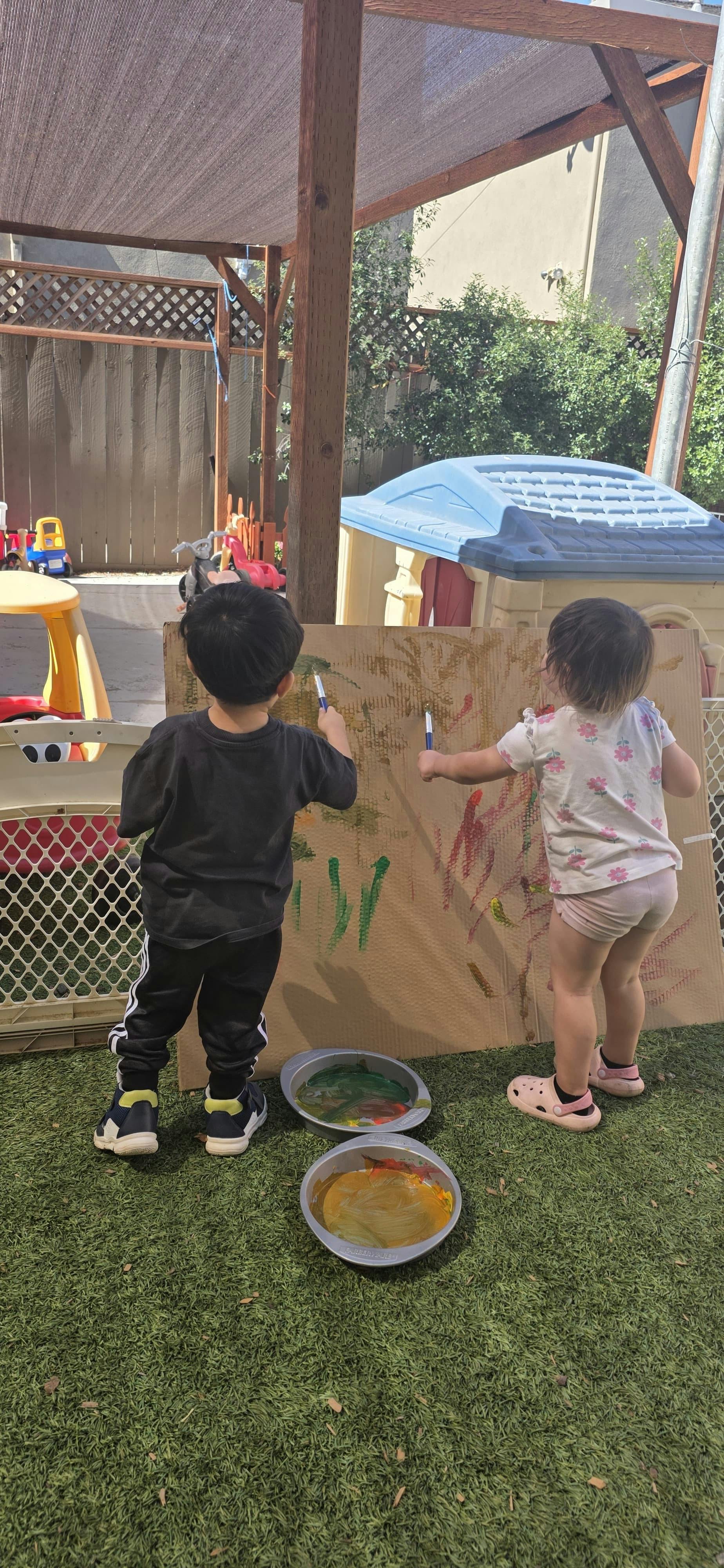 Two children painting on a large piece of cardboard outdoors