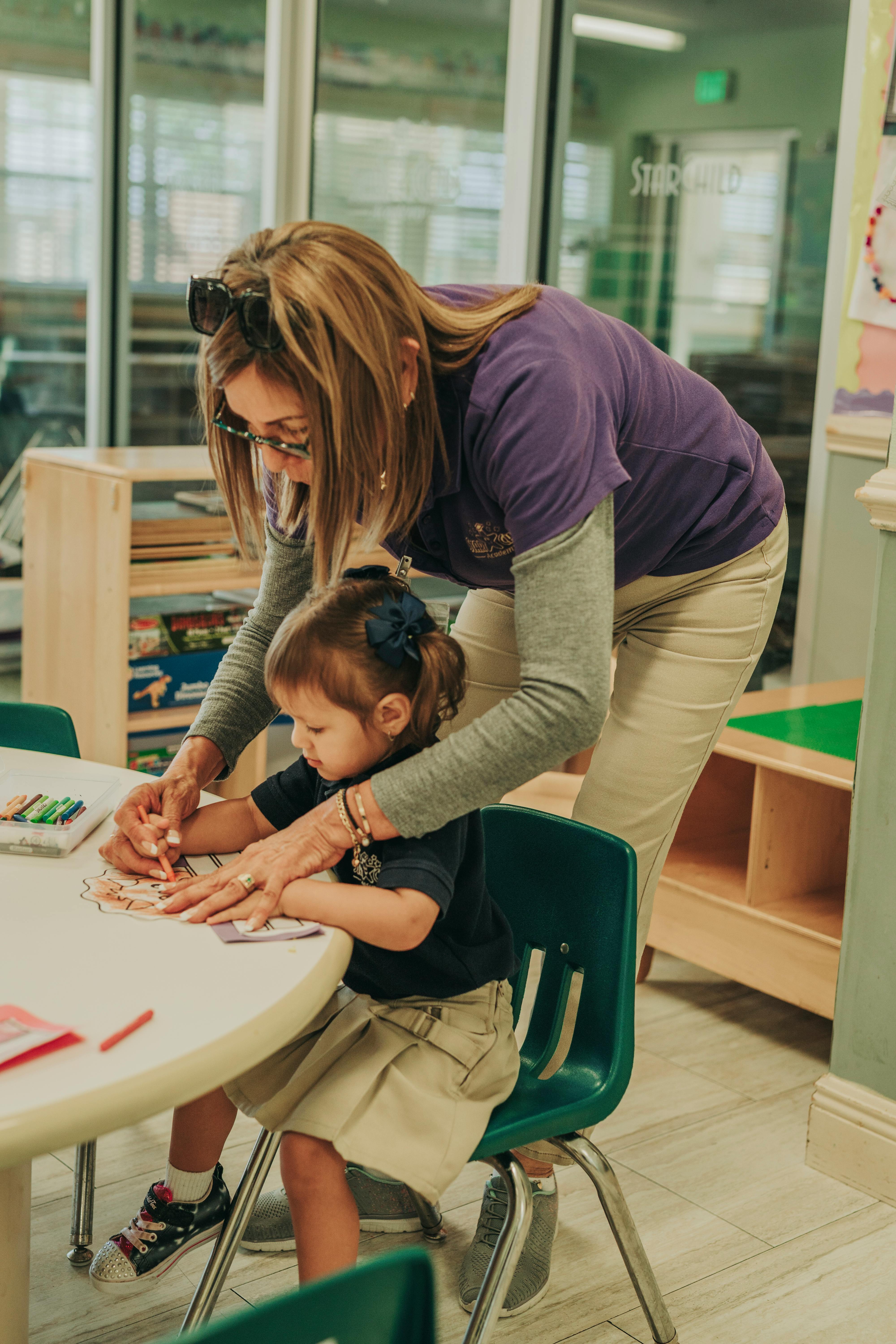 Person helping a child color at a table in a classroom