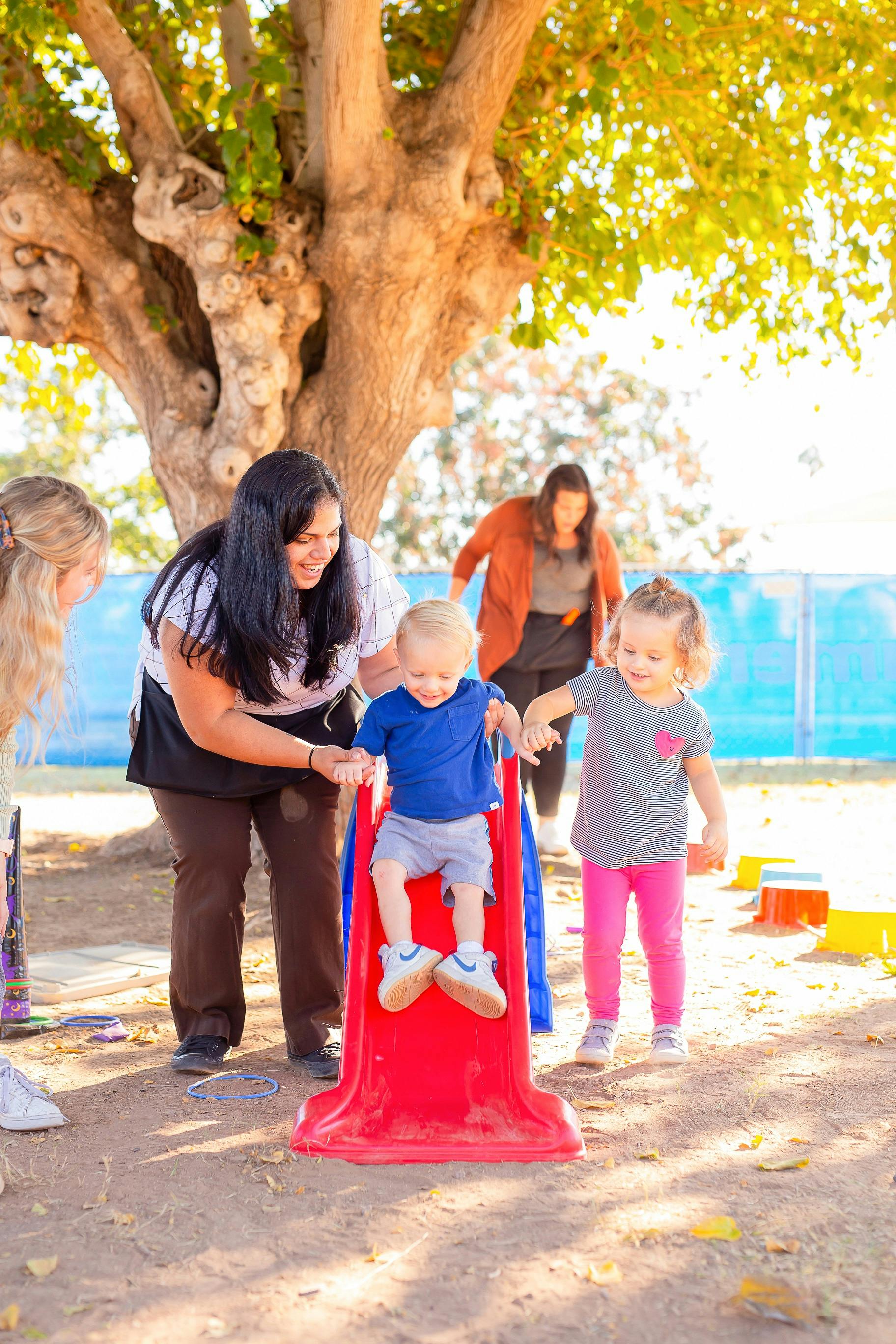 Adult assisting a child on a small slide while another child stands nearby