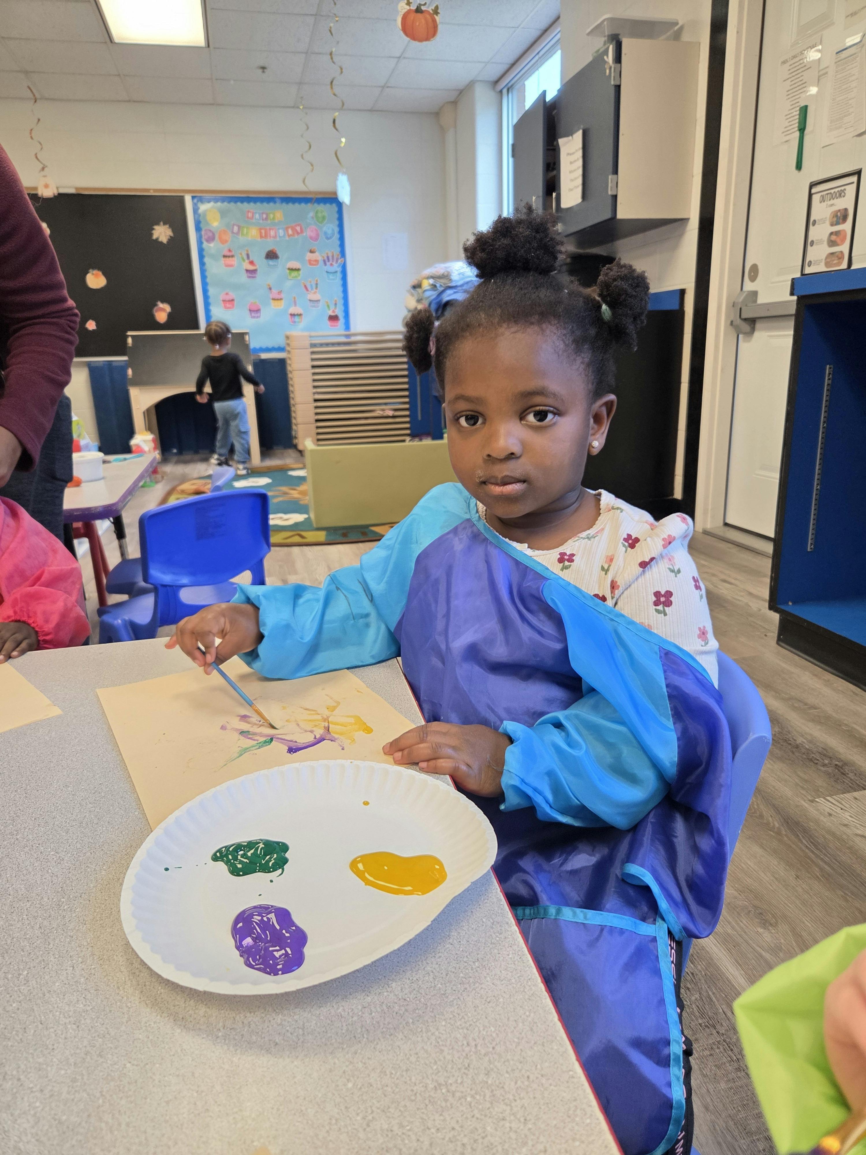 Child in smock painting on paper with green, yellow, and purple paints
