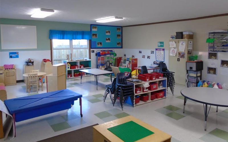 Empty classroom with small tables and chairs, shelves with toys, and whiteboards