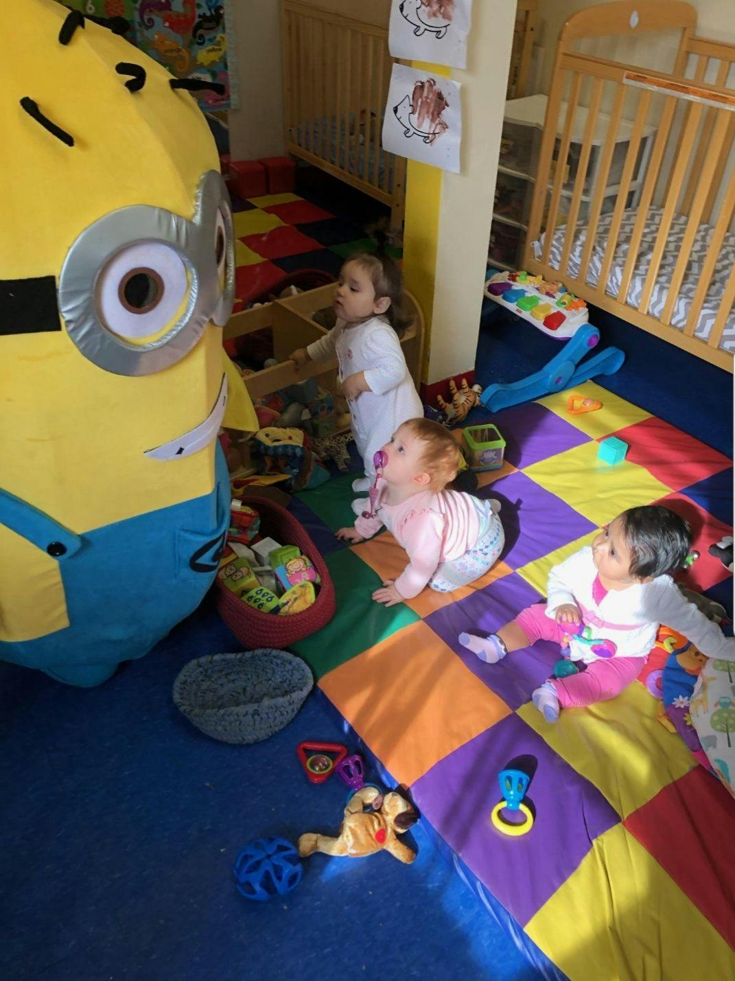 Three children playing on a colorful mat in a room with cribs
