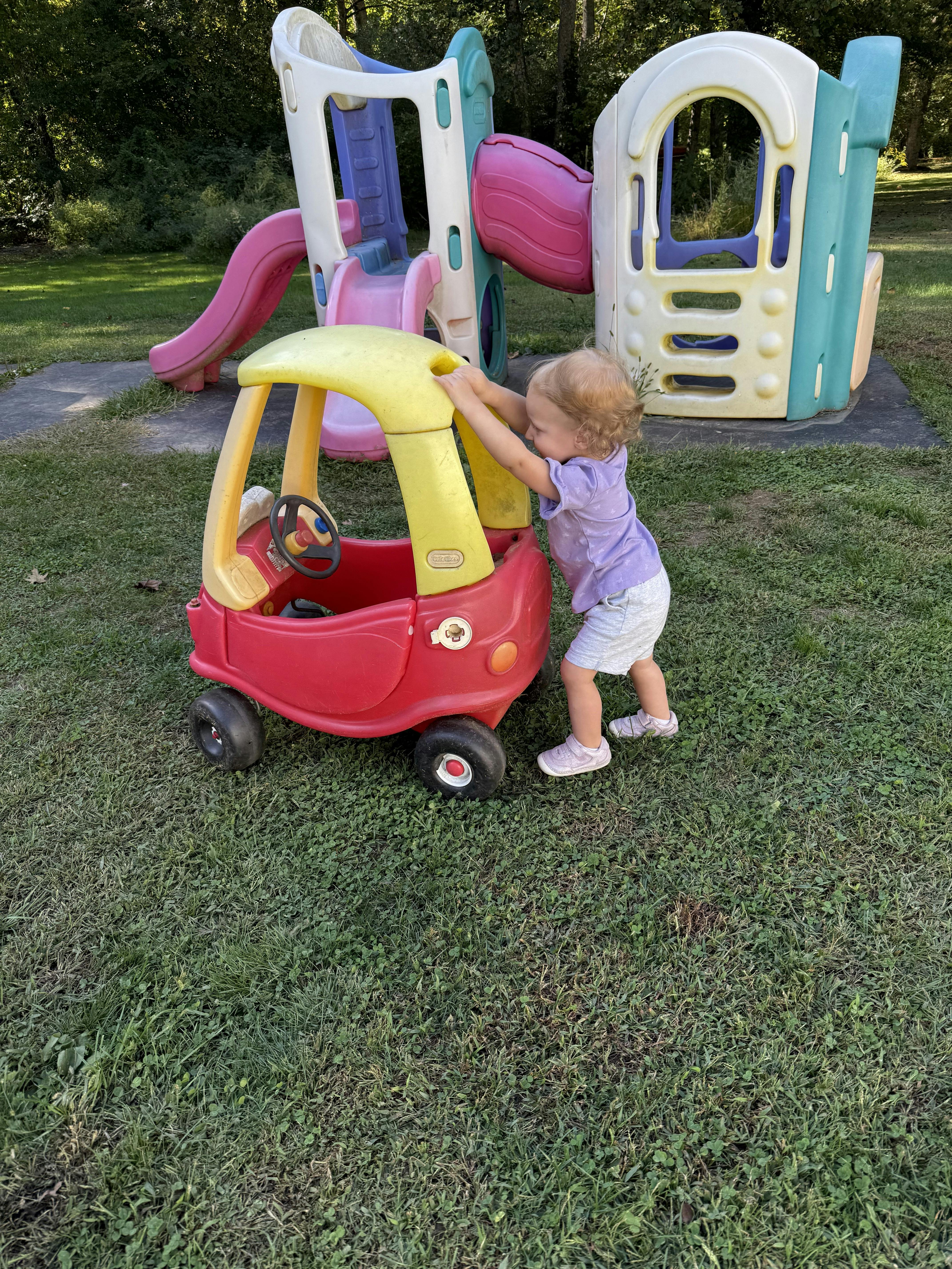 Child pushing a toy car on grass near a playground structure