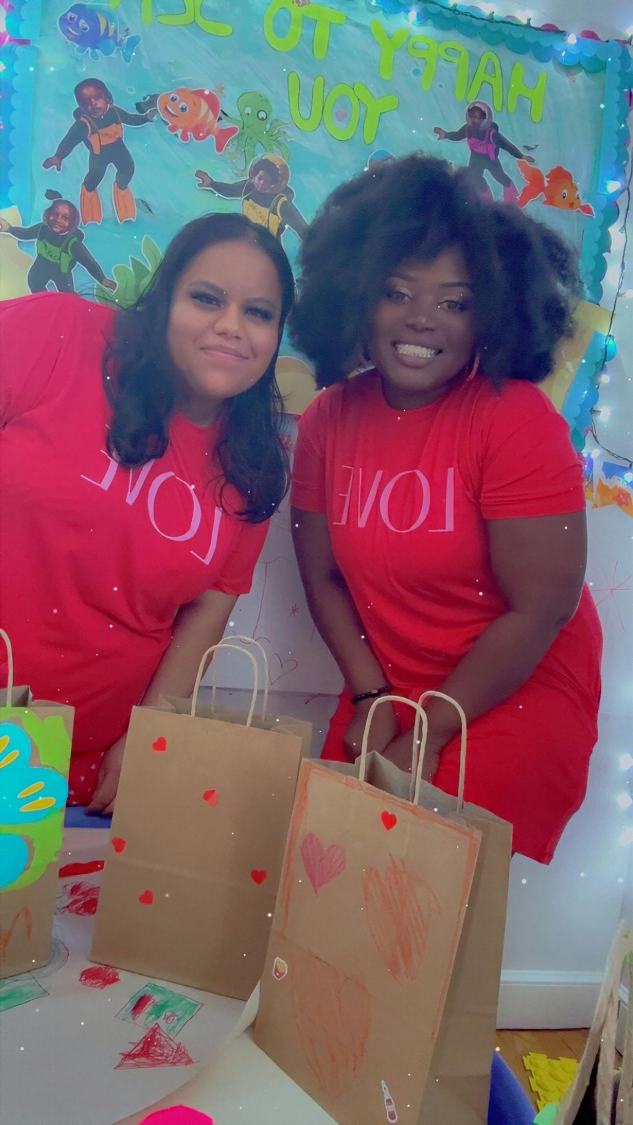 Two people wearing red "LOVE" shirts, standing behind paper bags with drawings