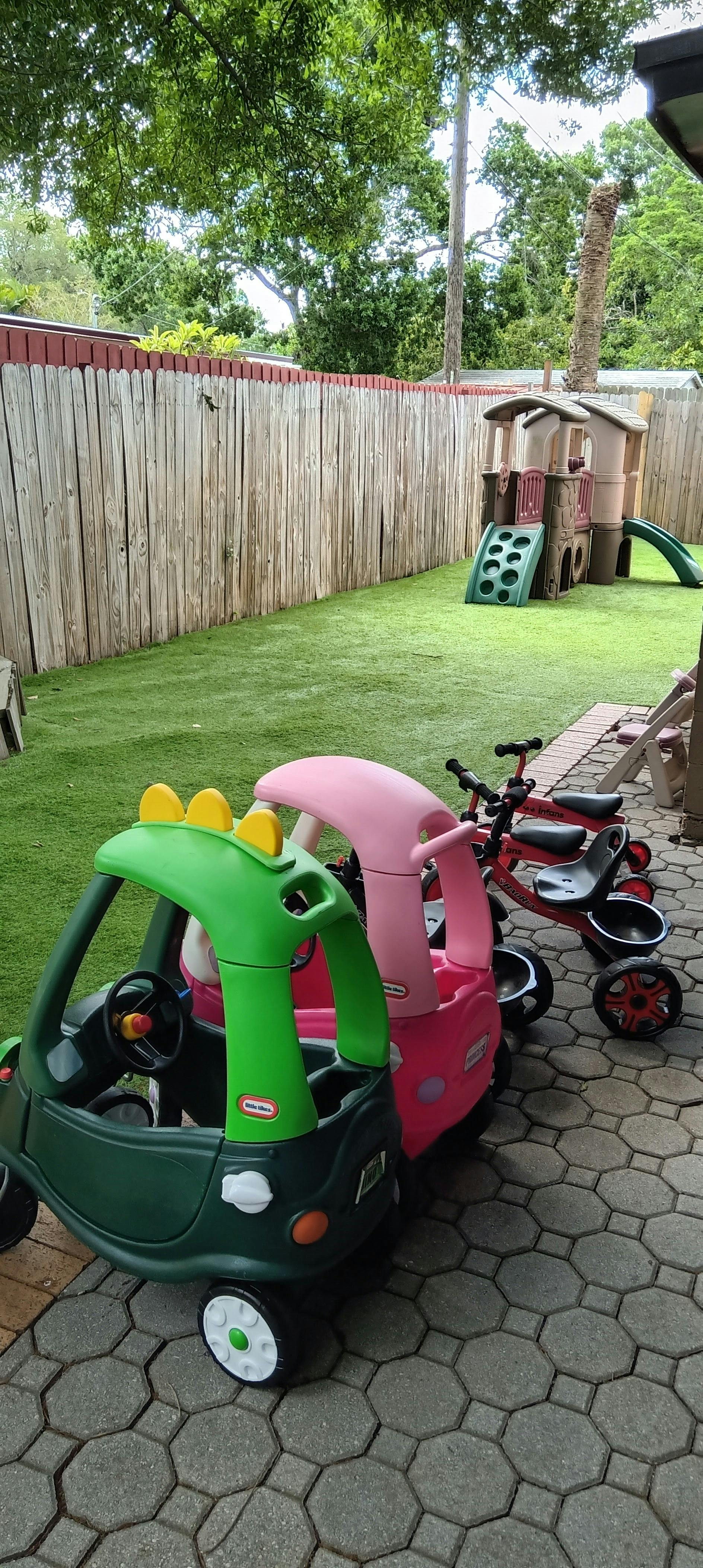 Plastic toy cars and tricycles on a paved patio next to a grassy yard