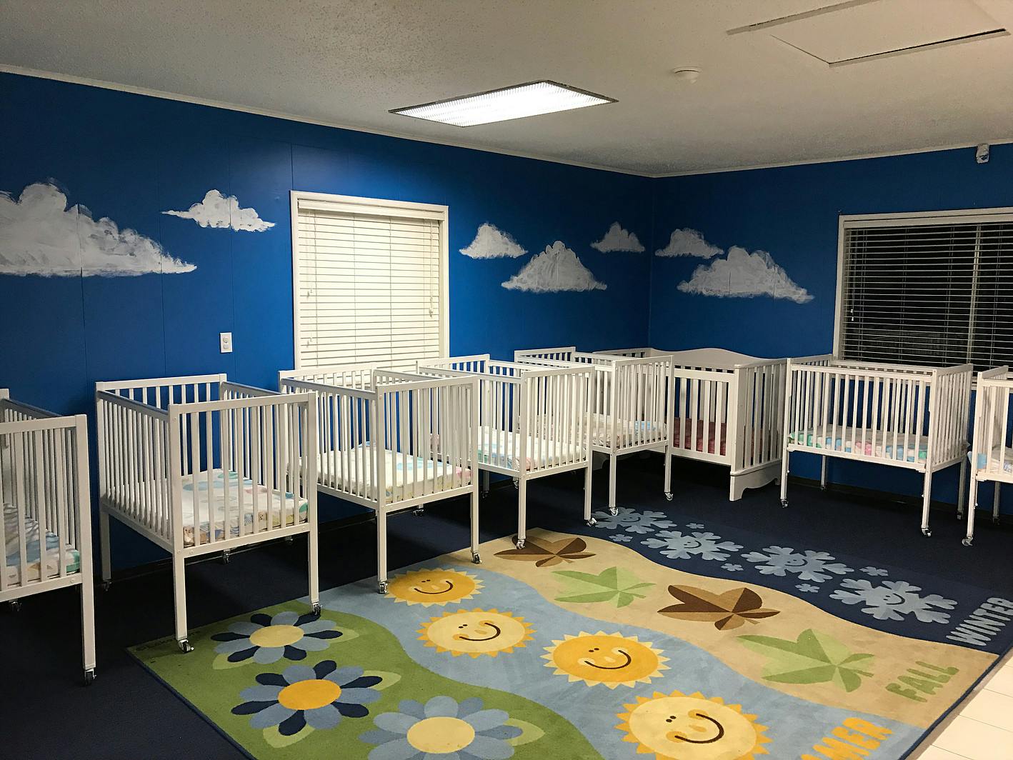 Room with multiple white cribs and a colorful rug