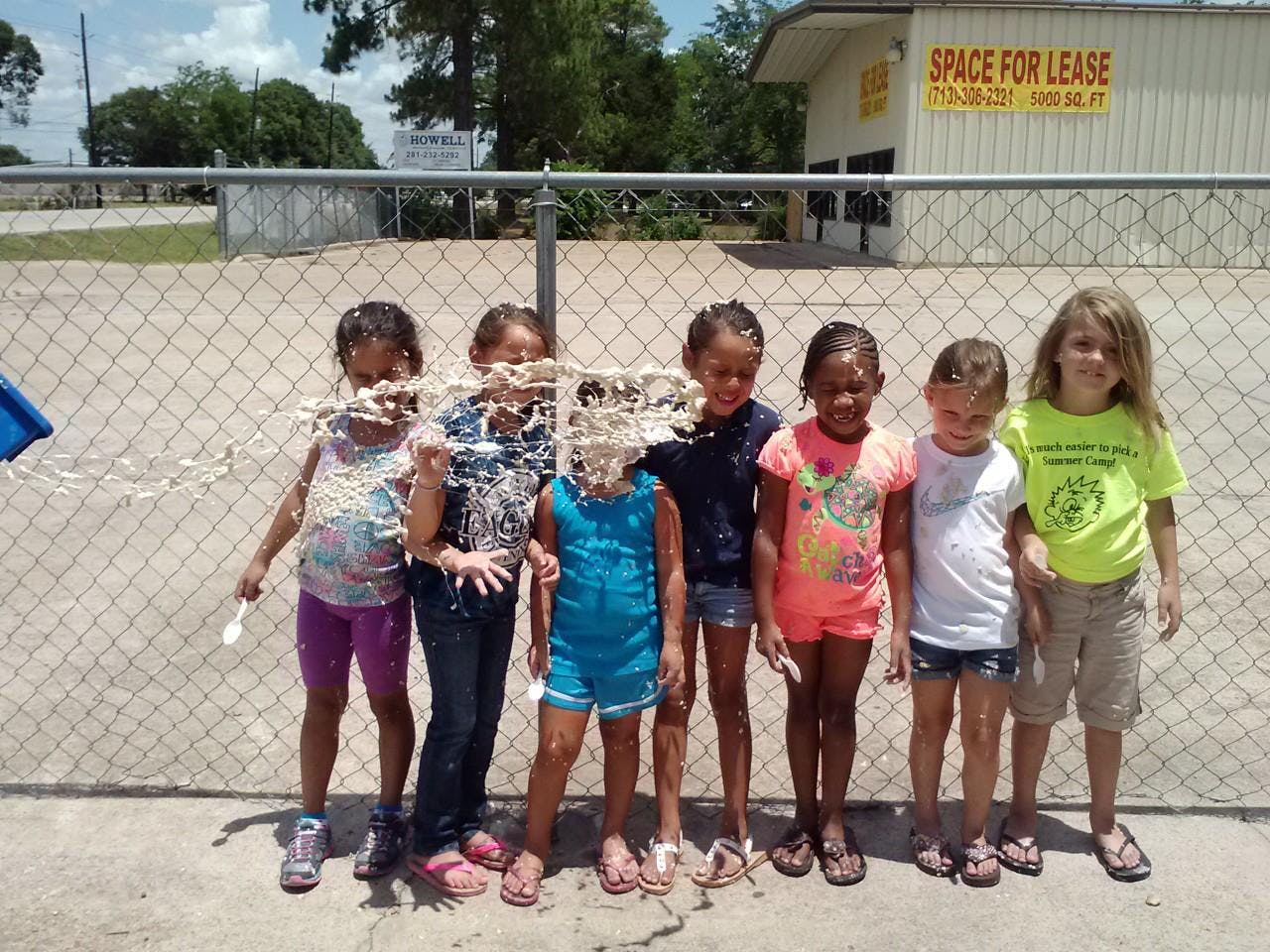 Six children standing in front of a fence, three with pie on their faces