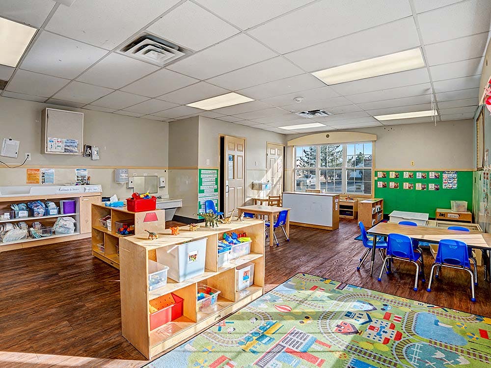 An empty children's classroom with small tables, chairs, shelves with toys, and a rug
