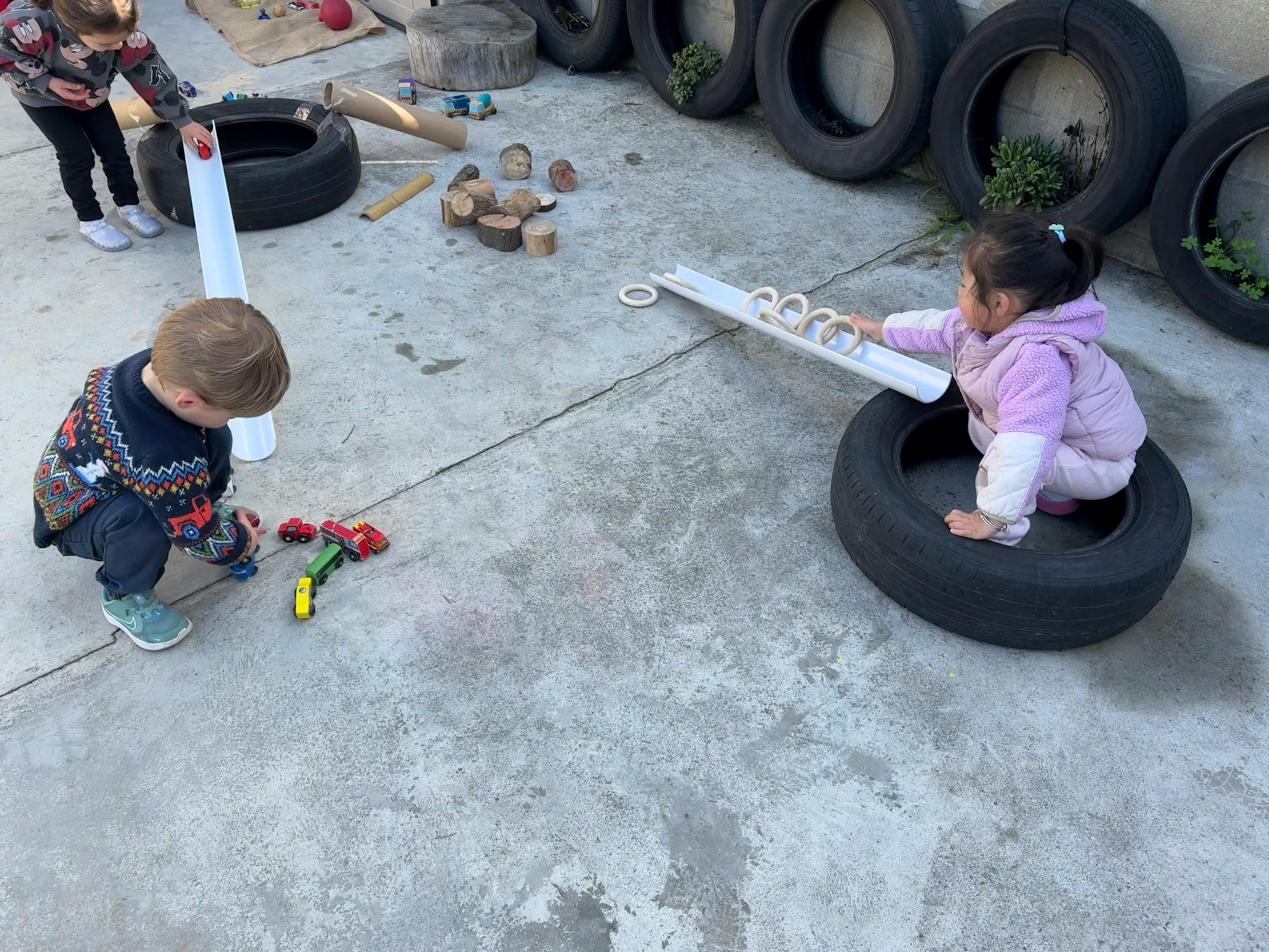 Three children playing outdoors with toy cars and a wooden ramp