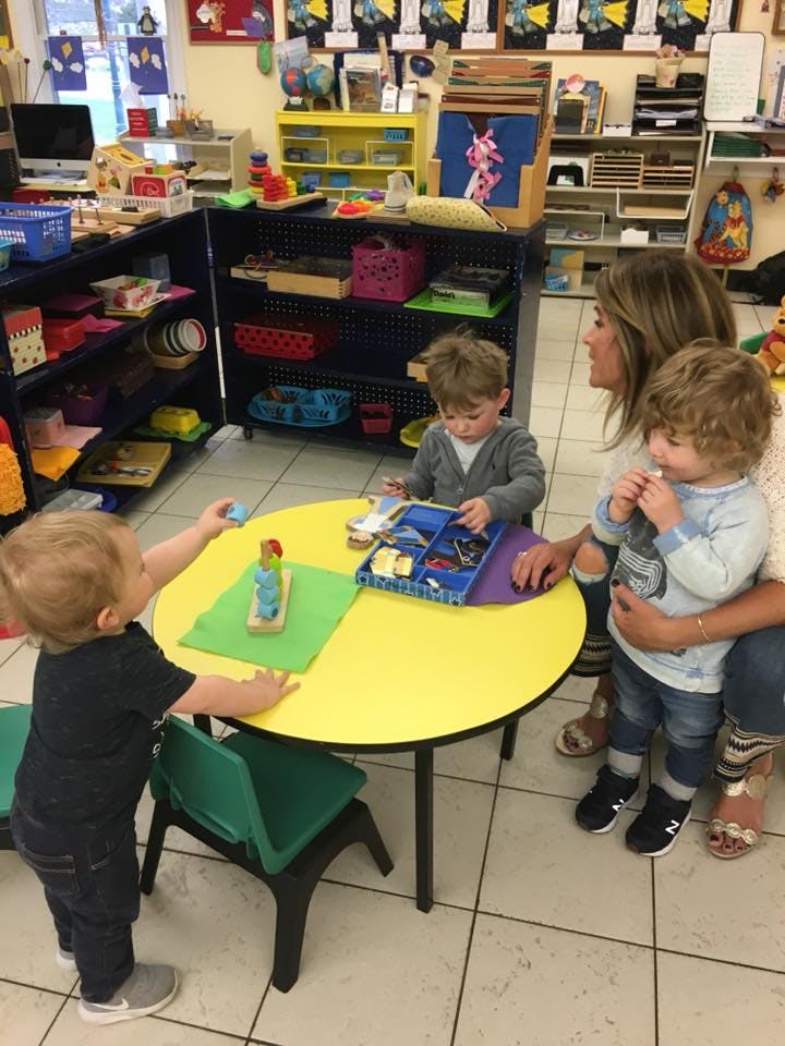 Adult and three children playing with toys at a yellow table in a classroom