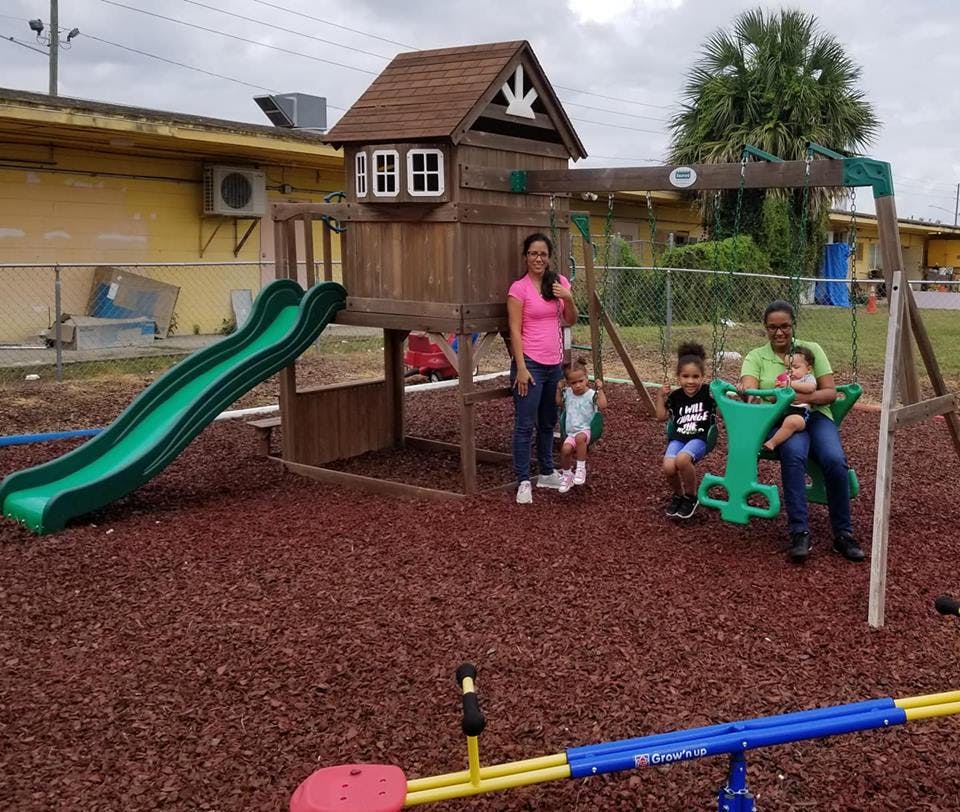 Two people and three children on a playground with a wooden playset and slide