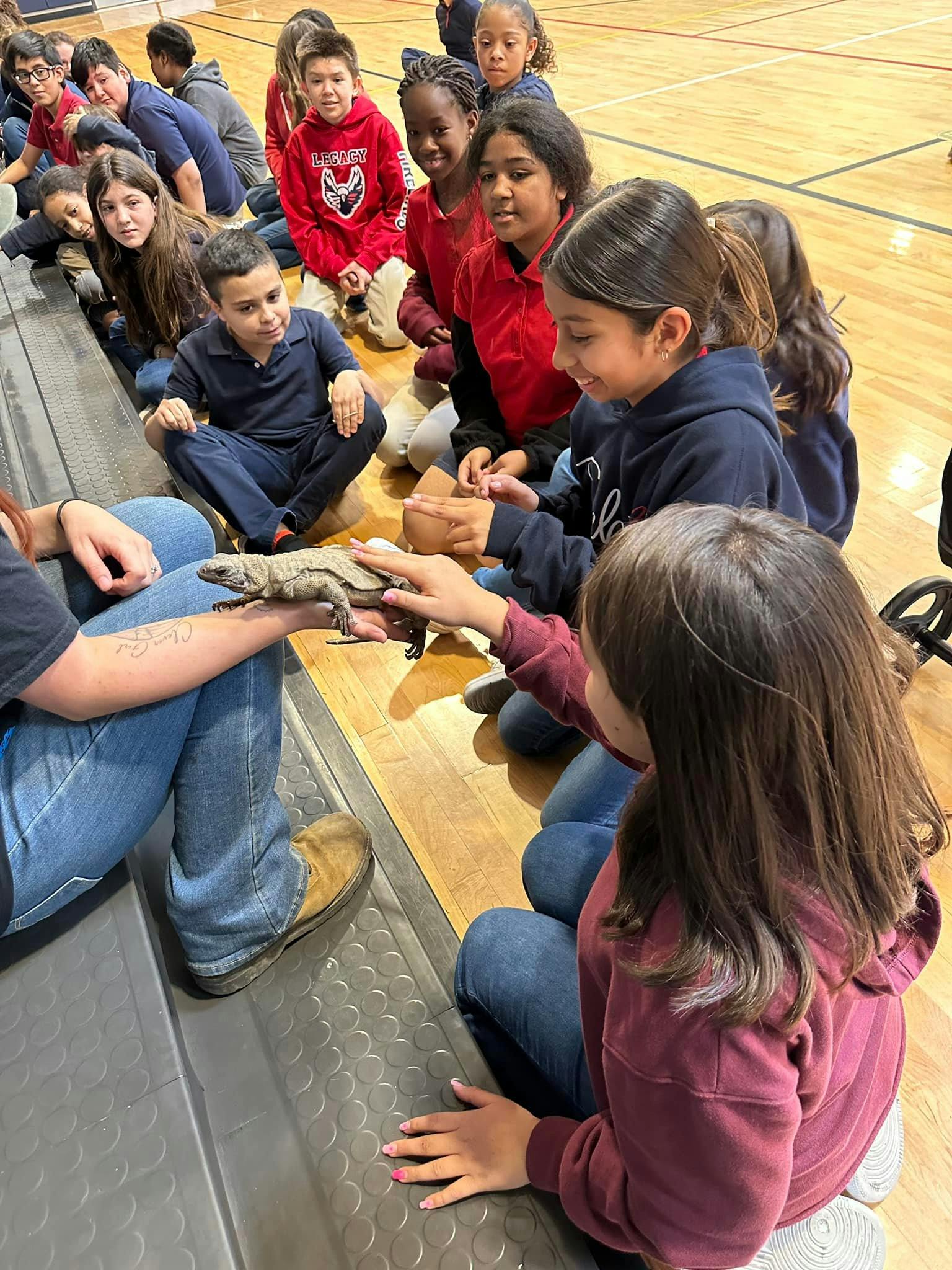 Children sitting on a gym floor looking at a lizard held by an adult