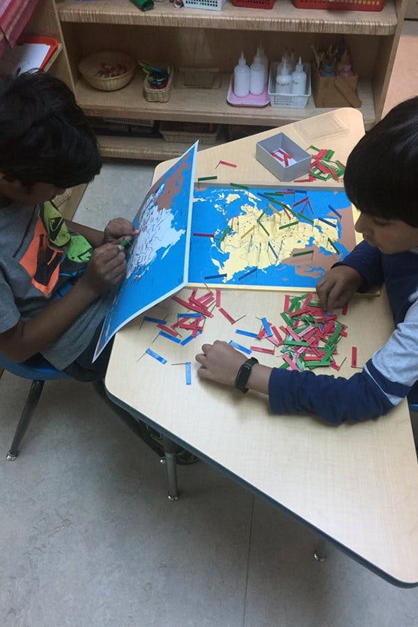 Two children working with a map puzzle and small colored pieces on a desk