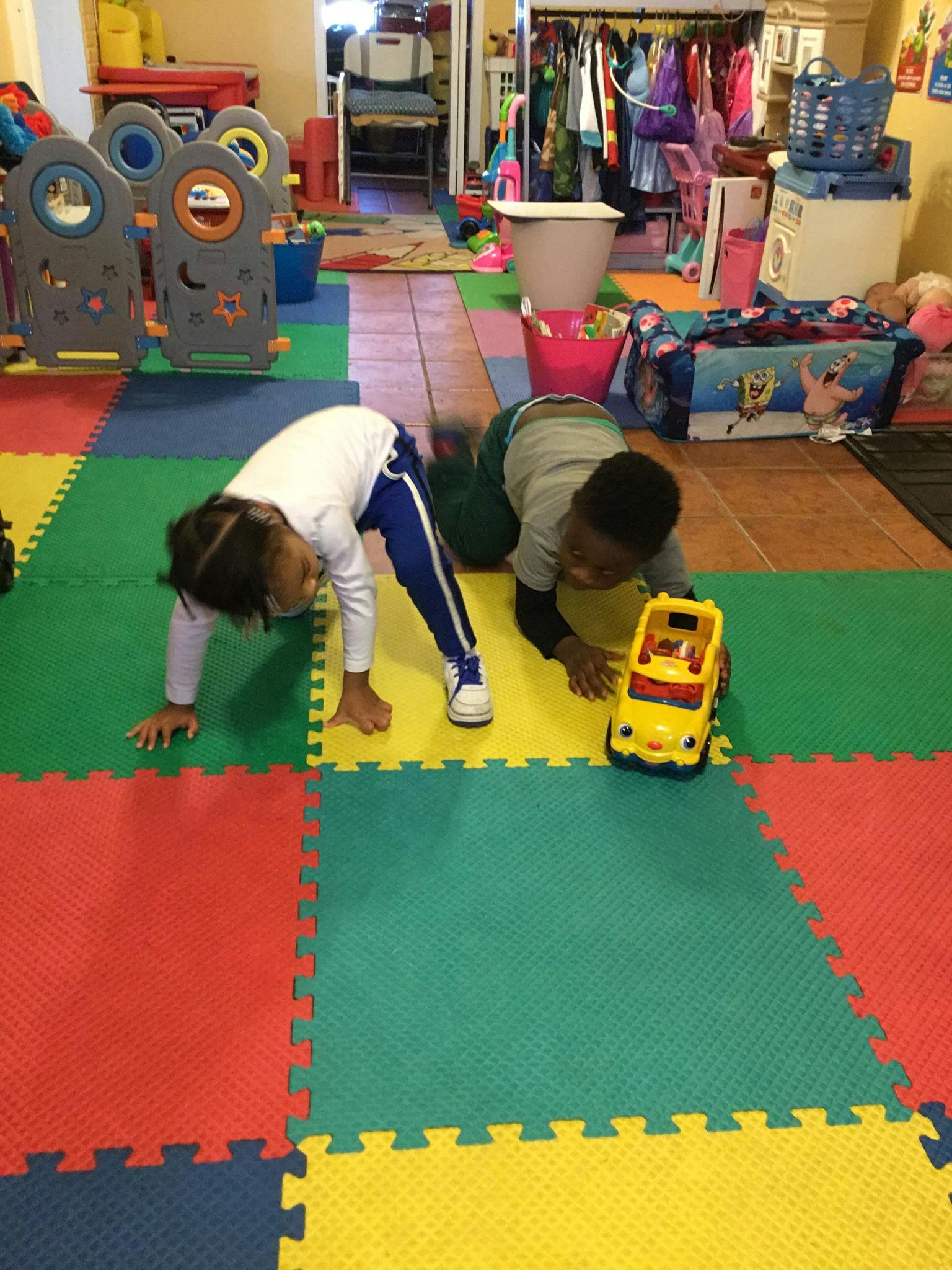 Two children playing on a colorful mat, one with a toy car