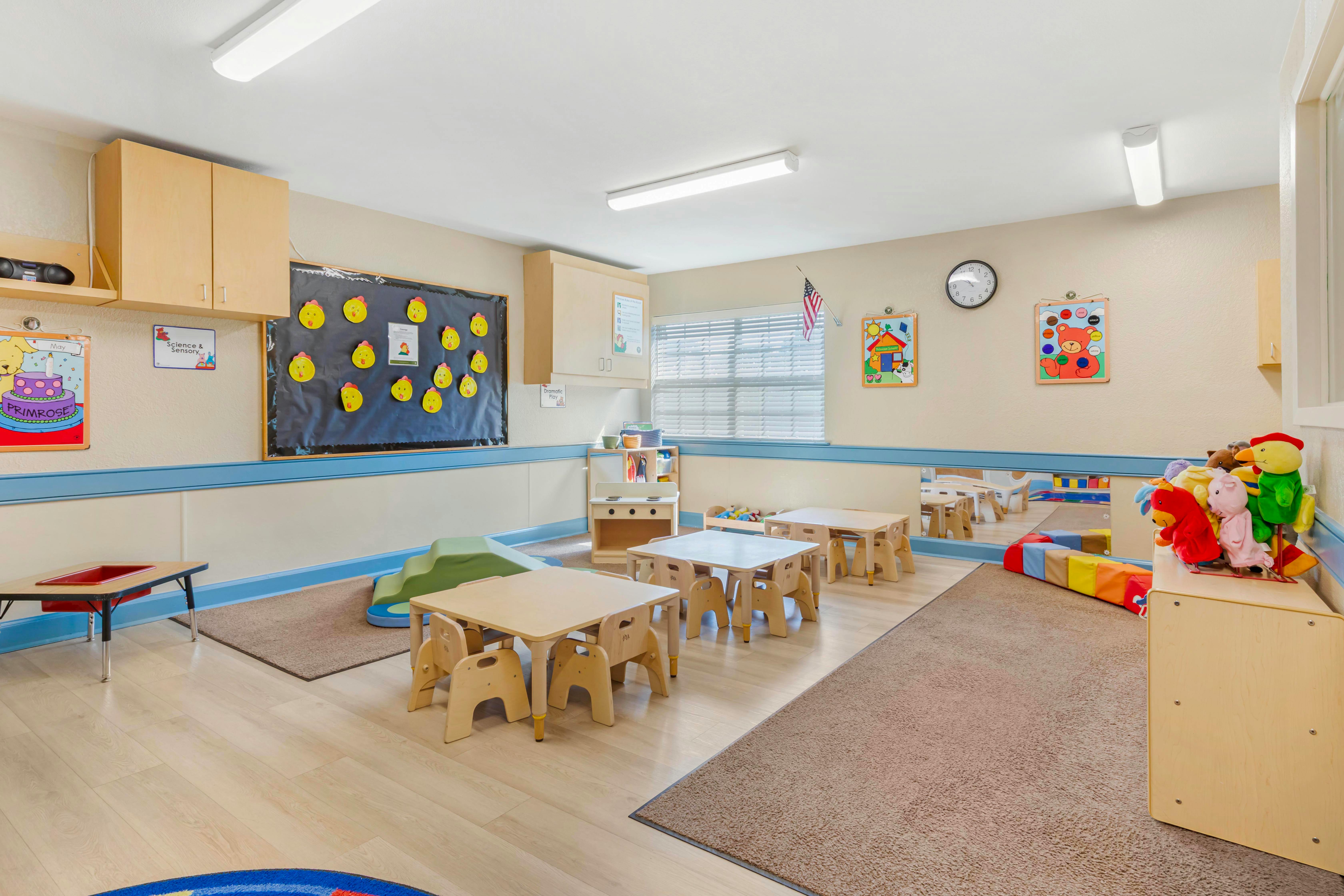 An empty preschool classroom with small tables and chairs, a play kitchen, and colorful decorations