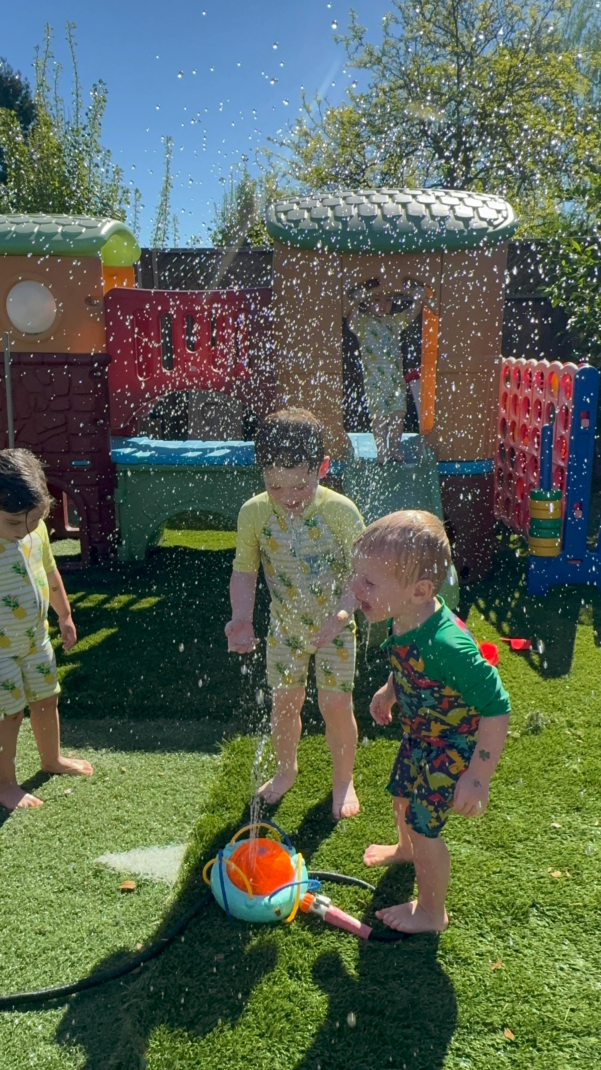 Three children playing in a sprinkler on green artificial turf