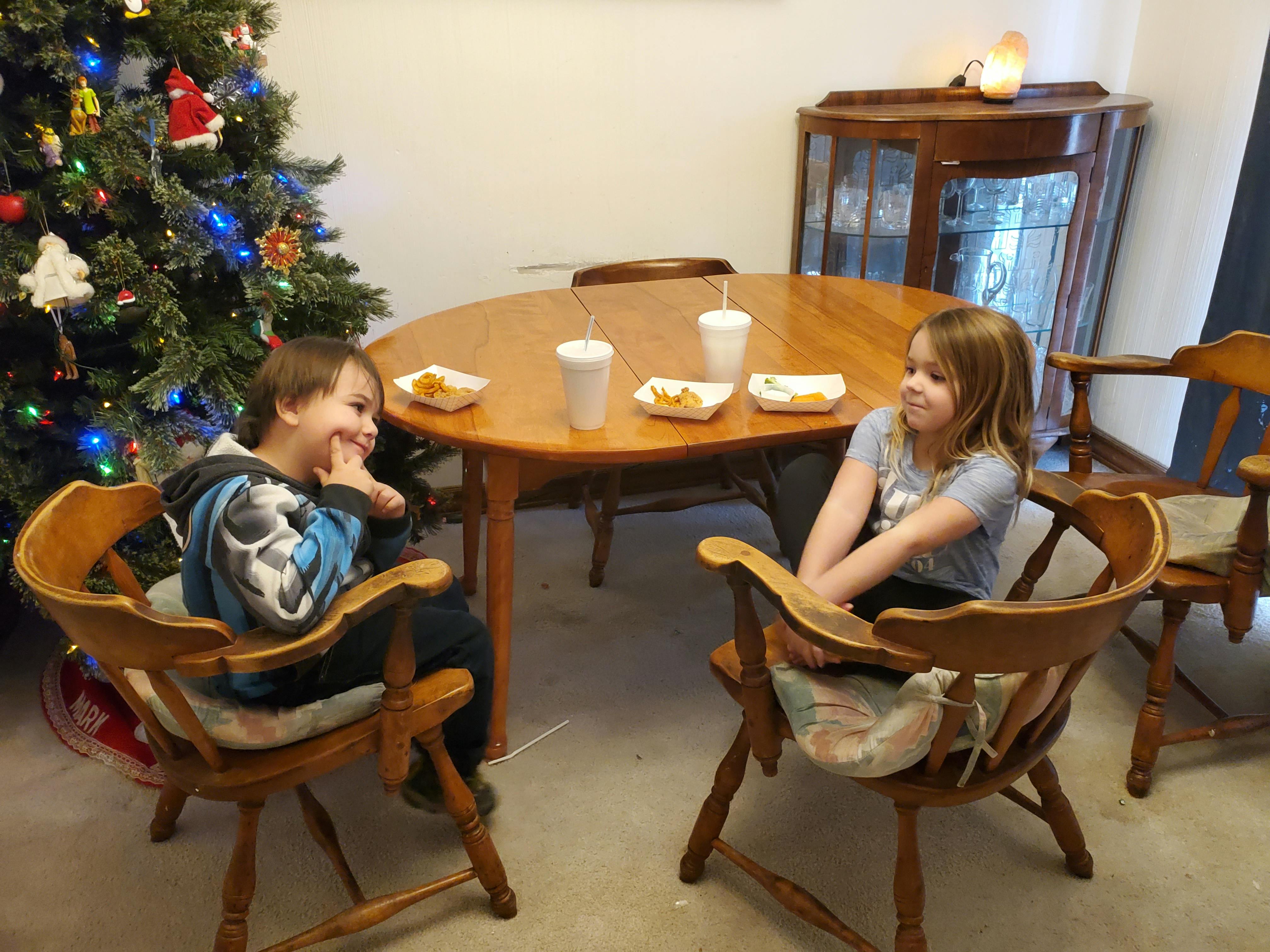 Two children sitting at a table with a Christmas tree nearby