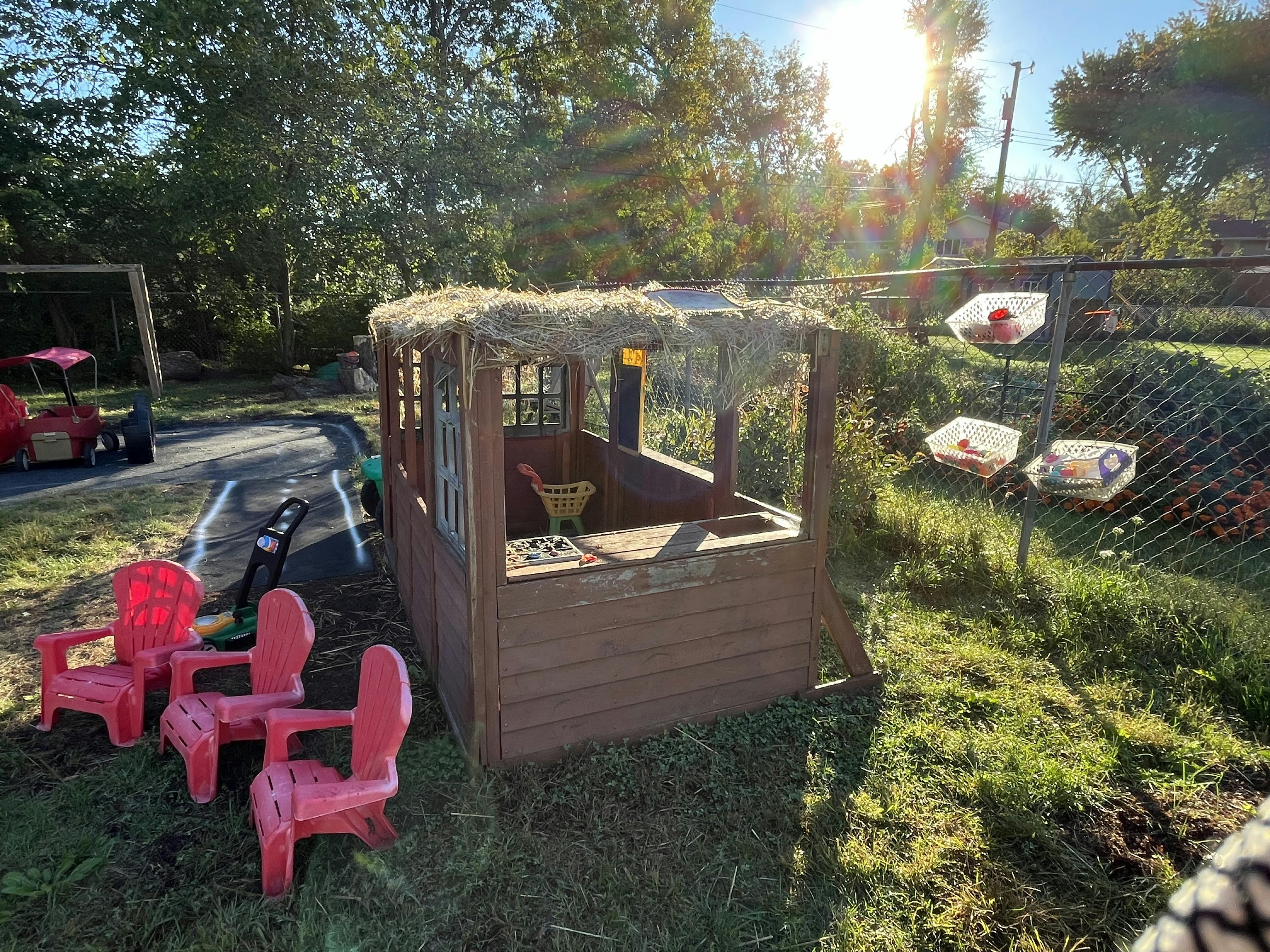 Wooden playhouse with a thatched roof and small plastic chairs in a yard