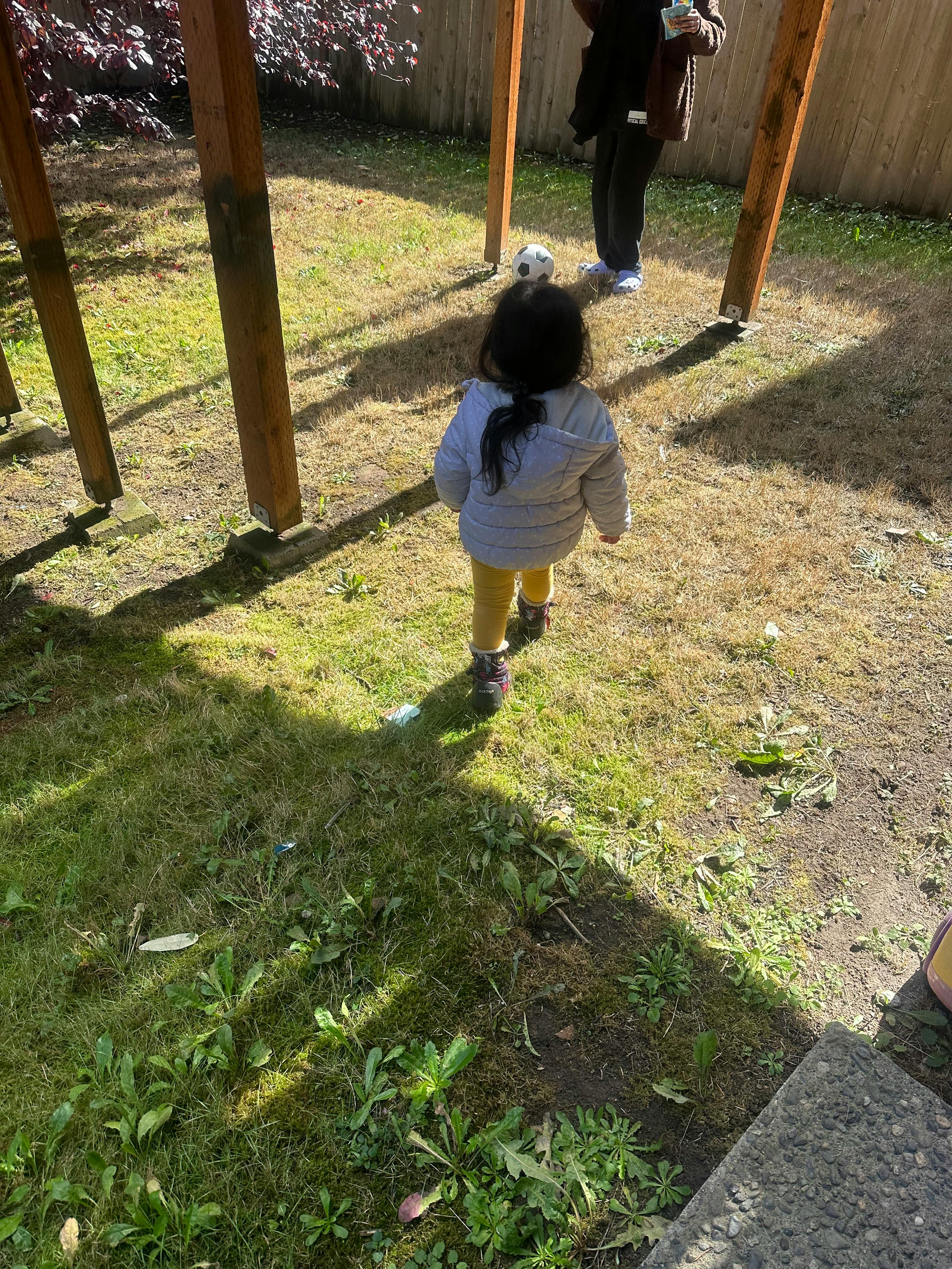 Child walking on grass toward a person standing under wooden beams
