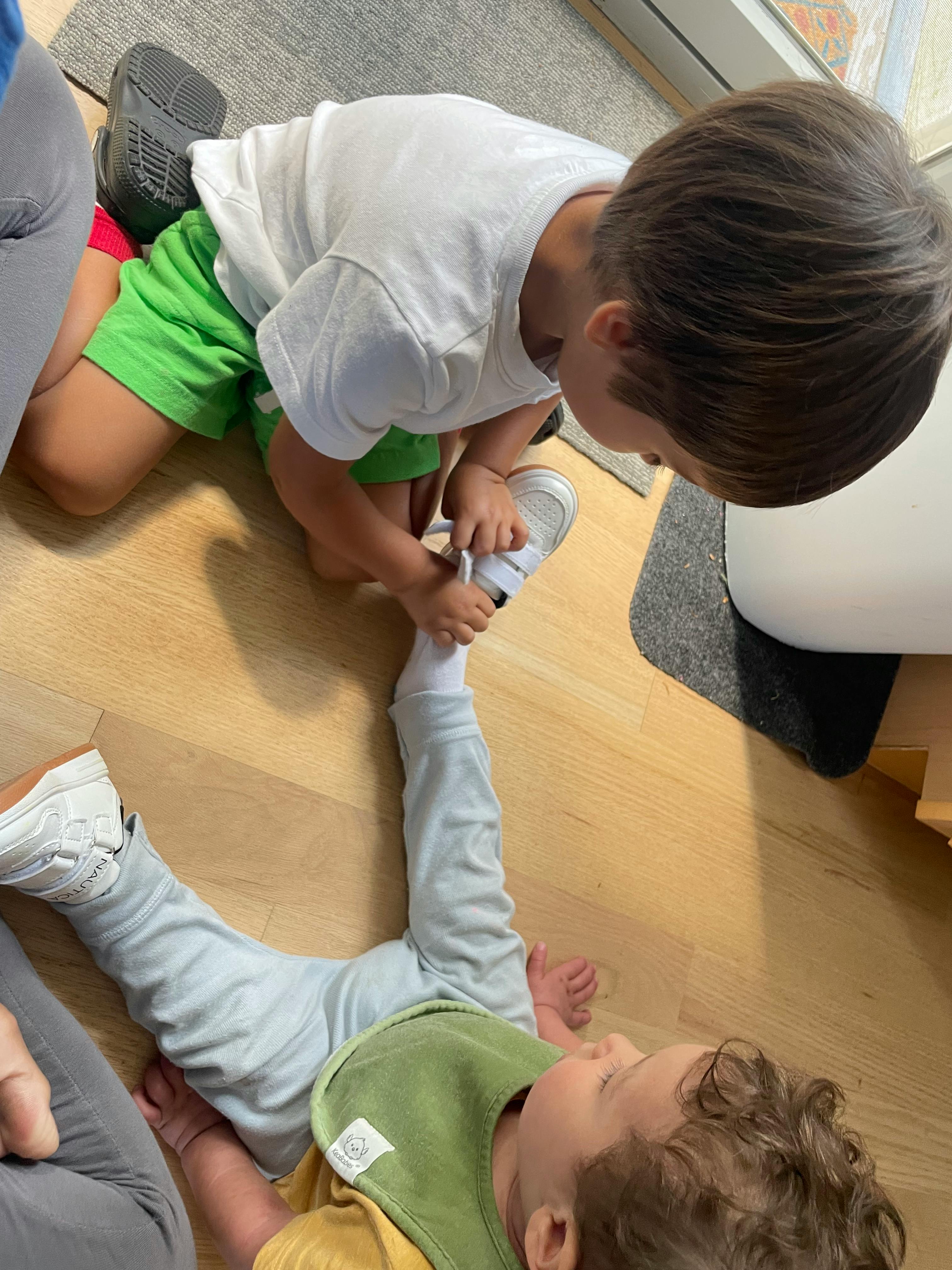Child kneeling to put a shoe on another child lying on the floor
