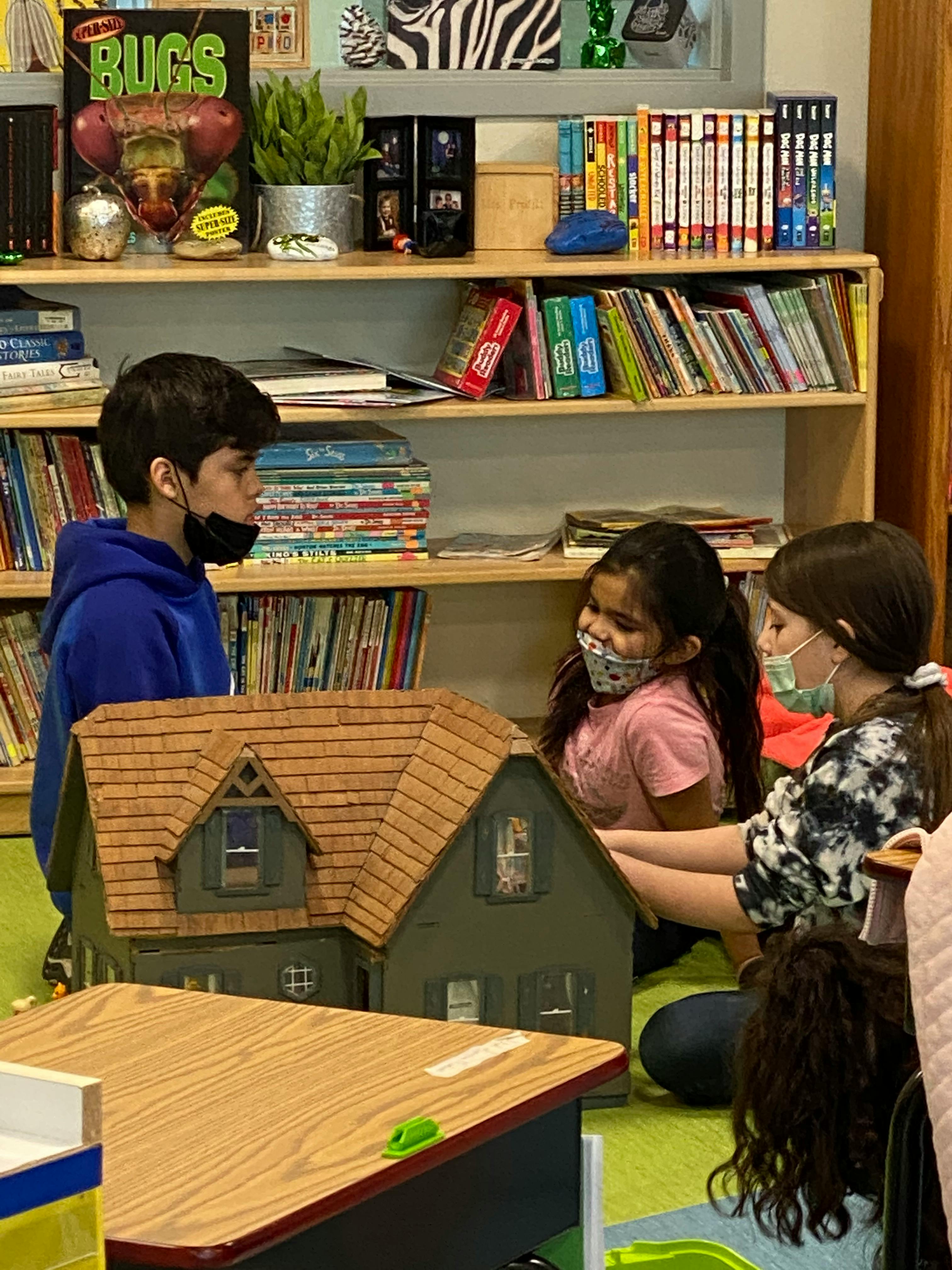Three children wearing masks looking at a dollhouse in a classroom