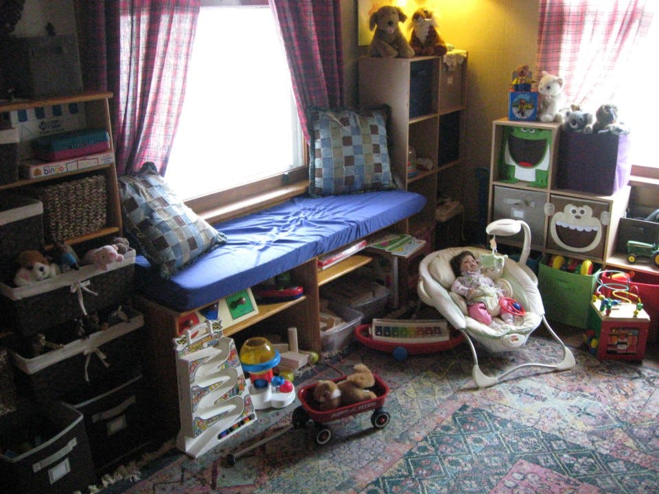 Child in a baby bouncer in a room with a window seat and shelves of toys