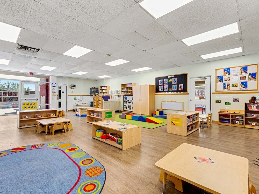 Empty classroom with small tables and chairs, shelves with toys, and colorful rugs