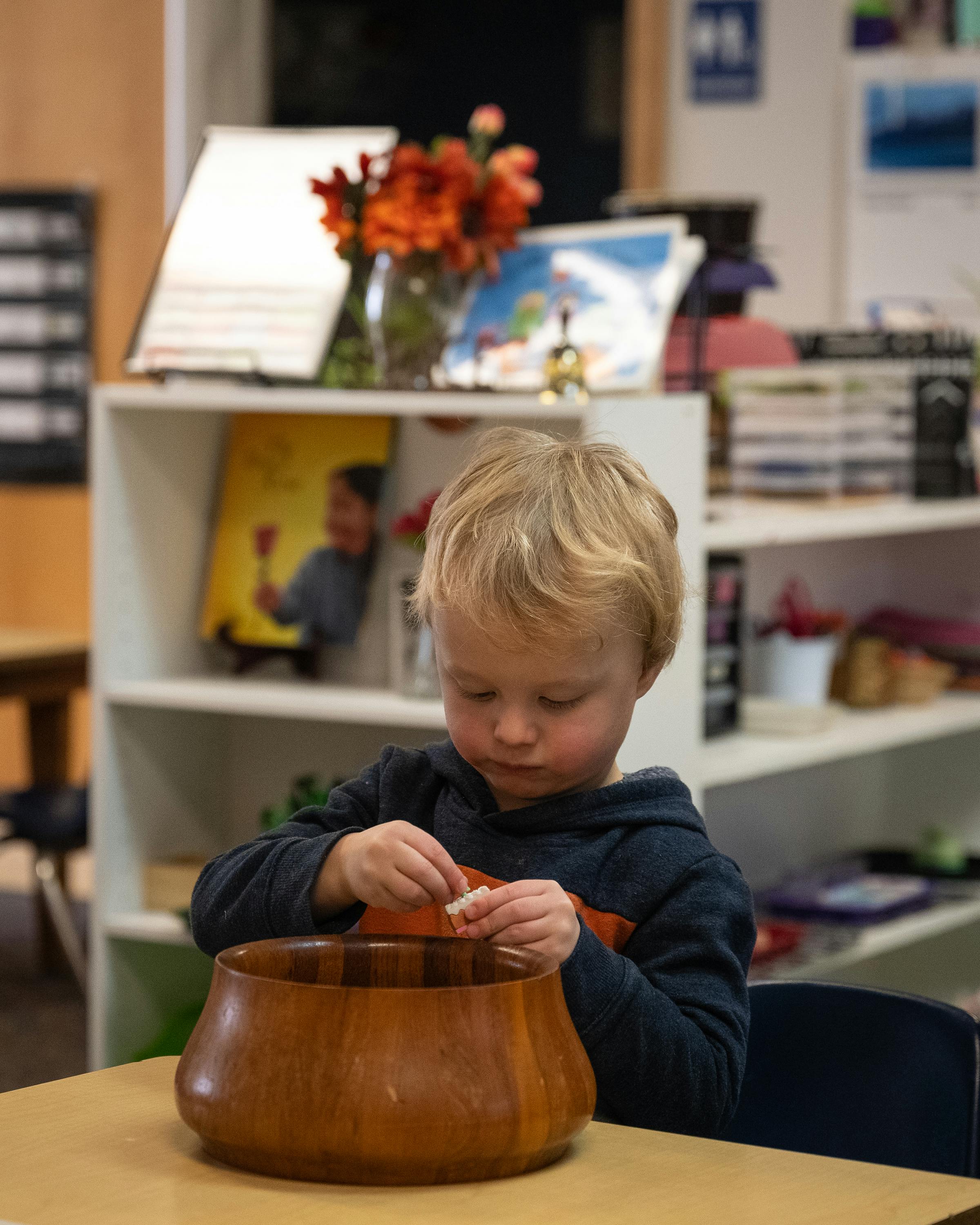 Child sitting at a table, holding a small object over a wooden bowl