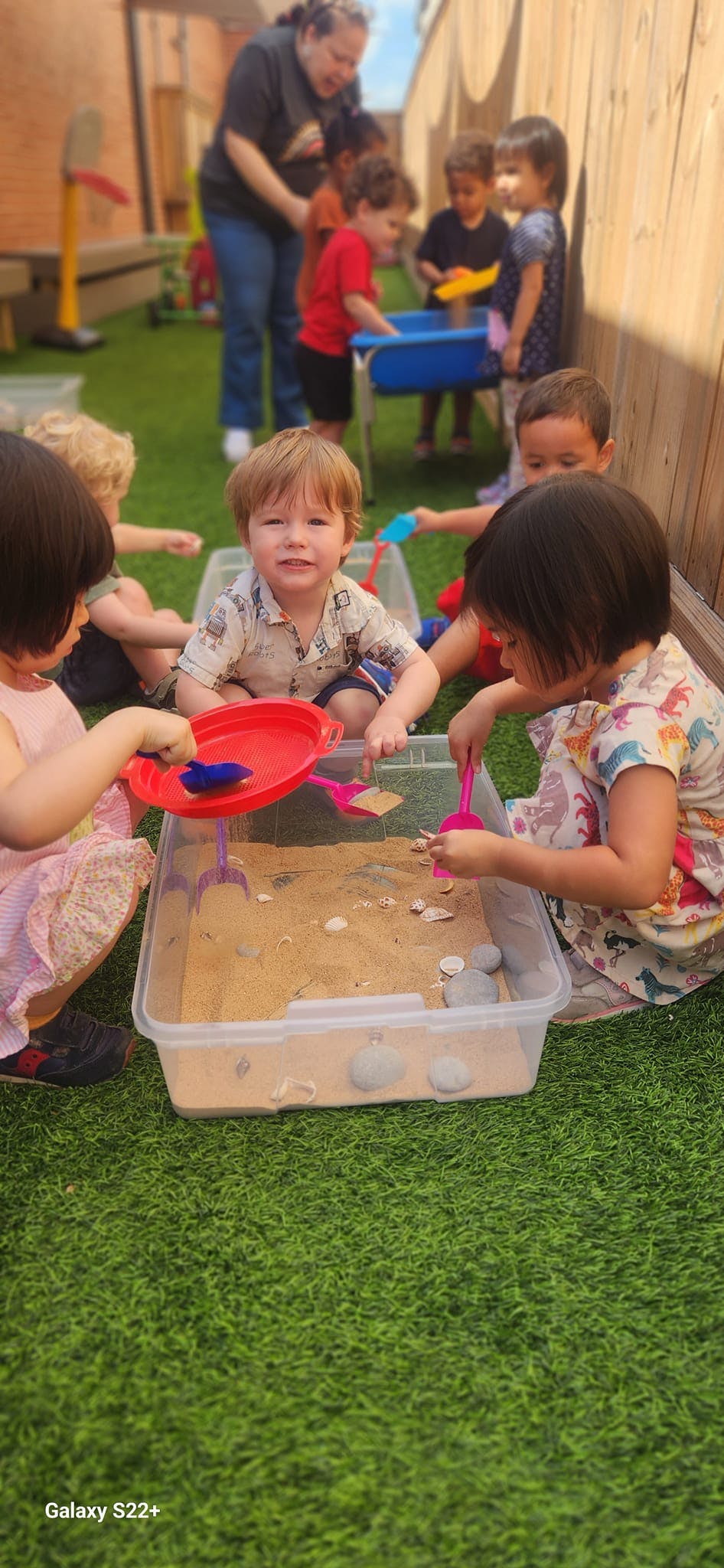 Children playing with sand and toys in a container on artificial grass