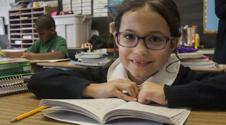 Child in glasses looking up from an open book at a desk in a classroom