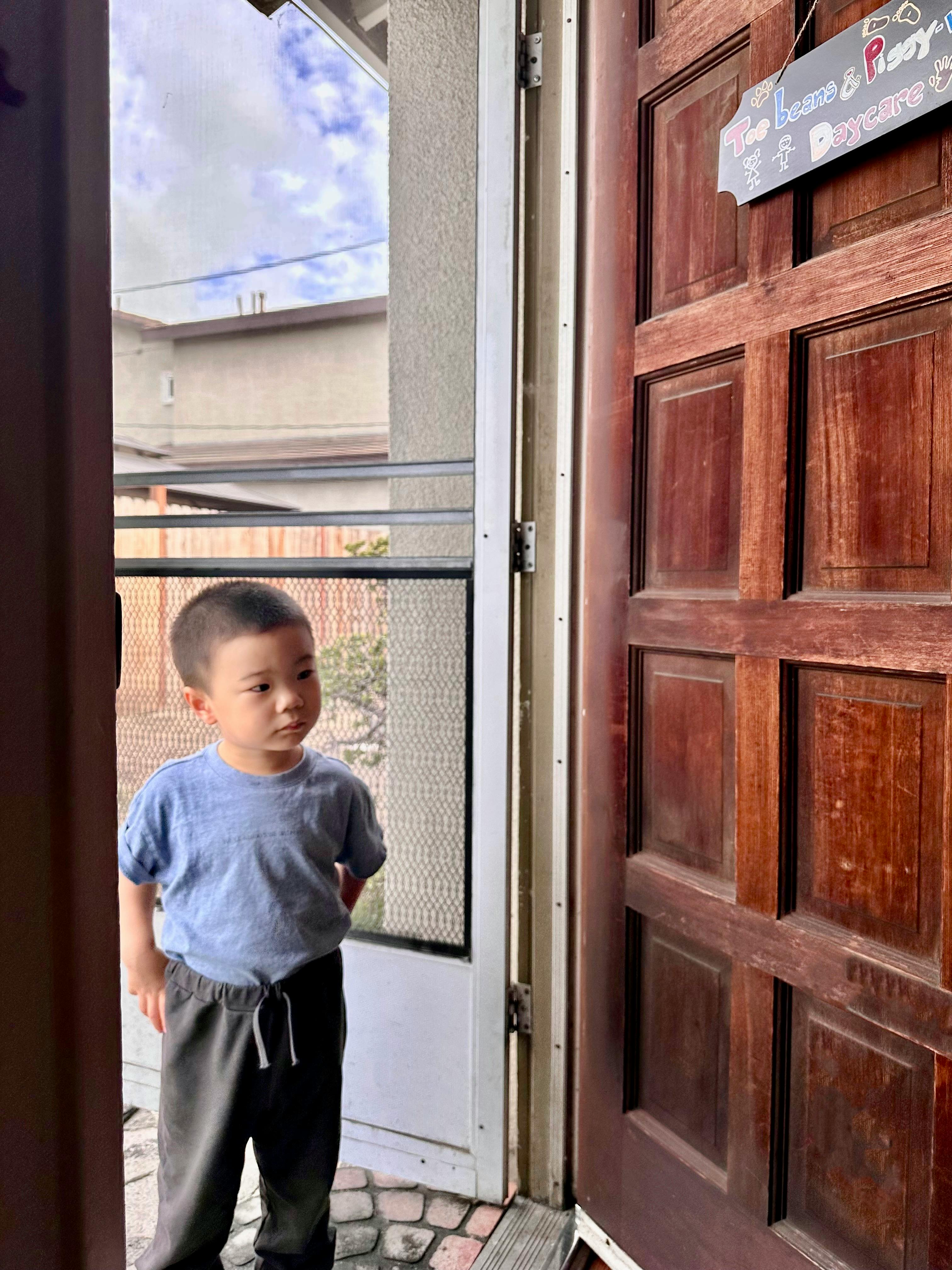 Child standing in an open doorway with a daycare sign hanging on the wood door