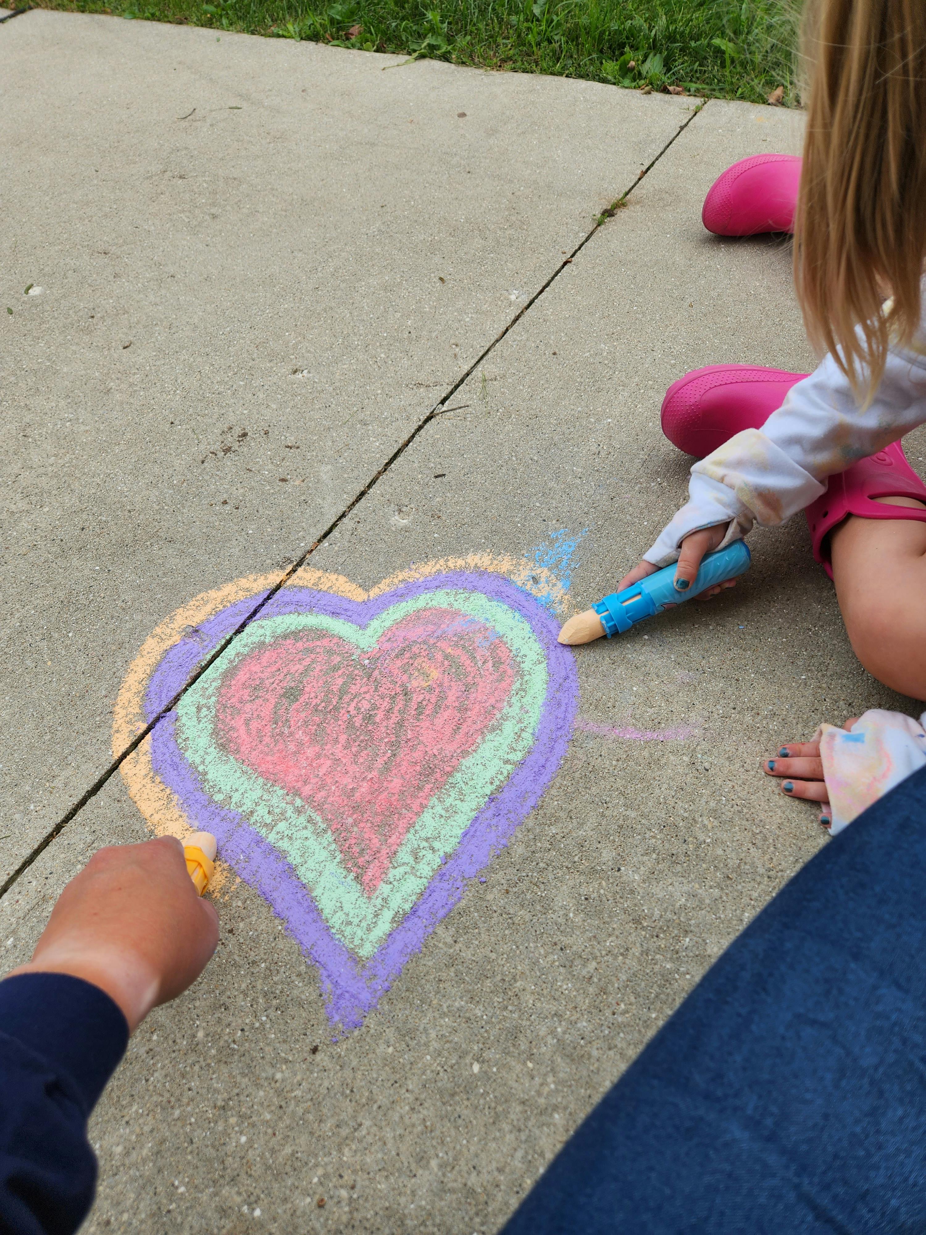 Two people drawing a multi-colored heart with chalk on concrete