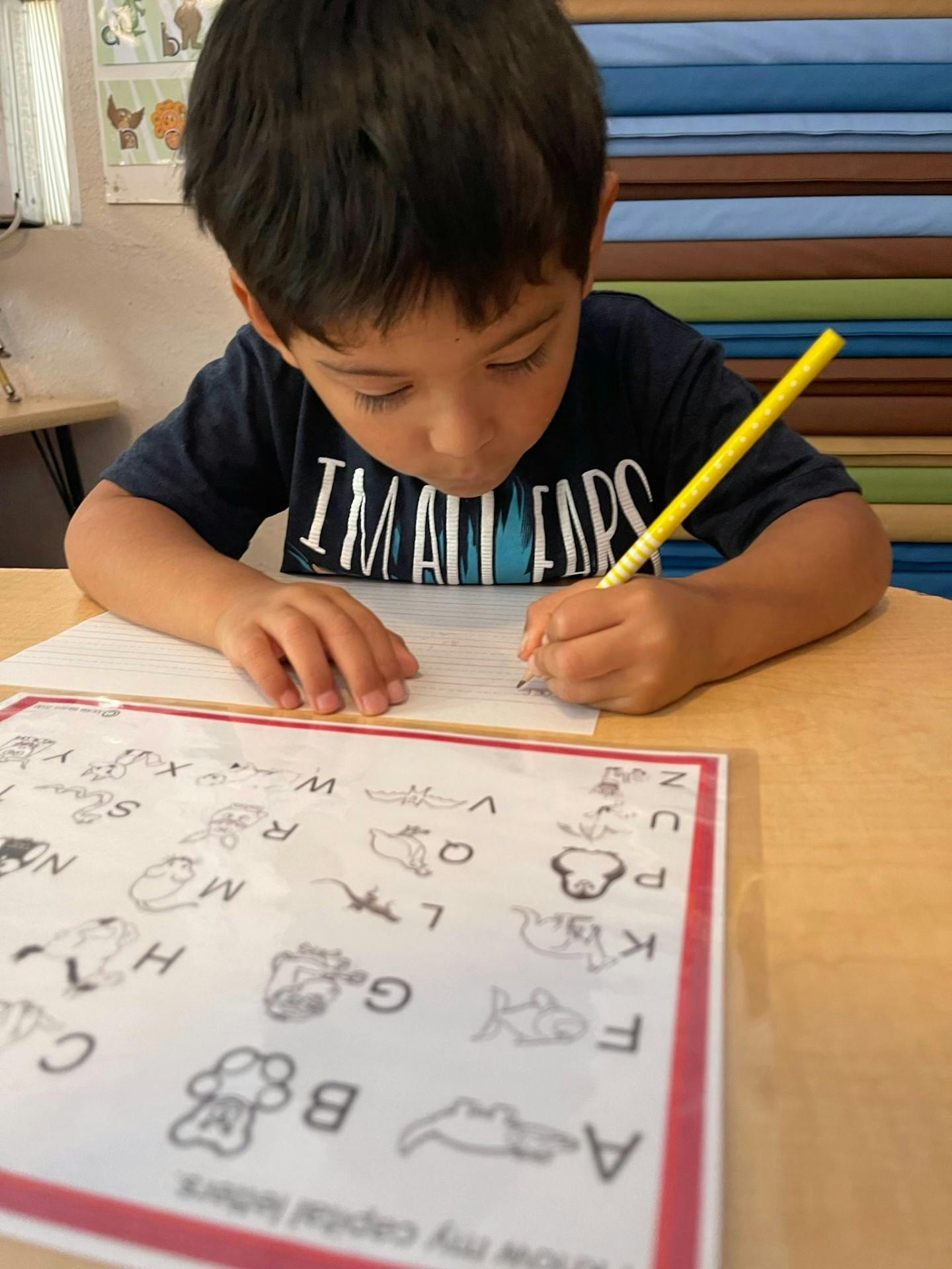 Child writing on paper with a pencil, an alphabet sheet on the table