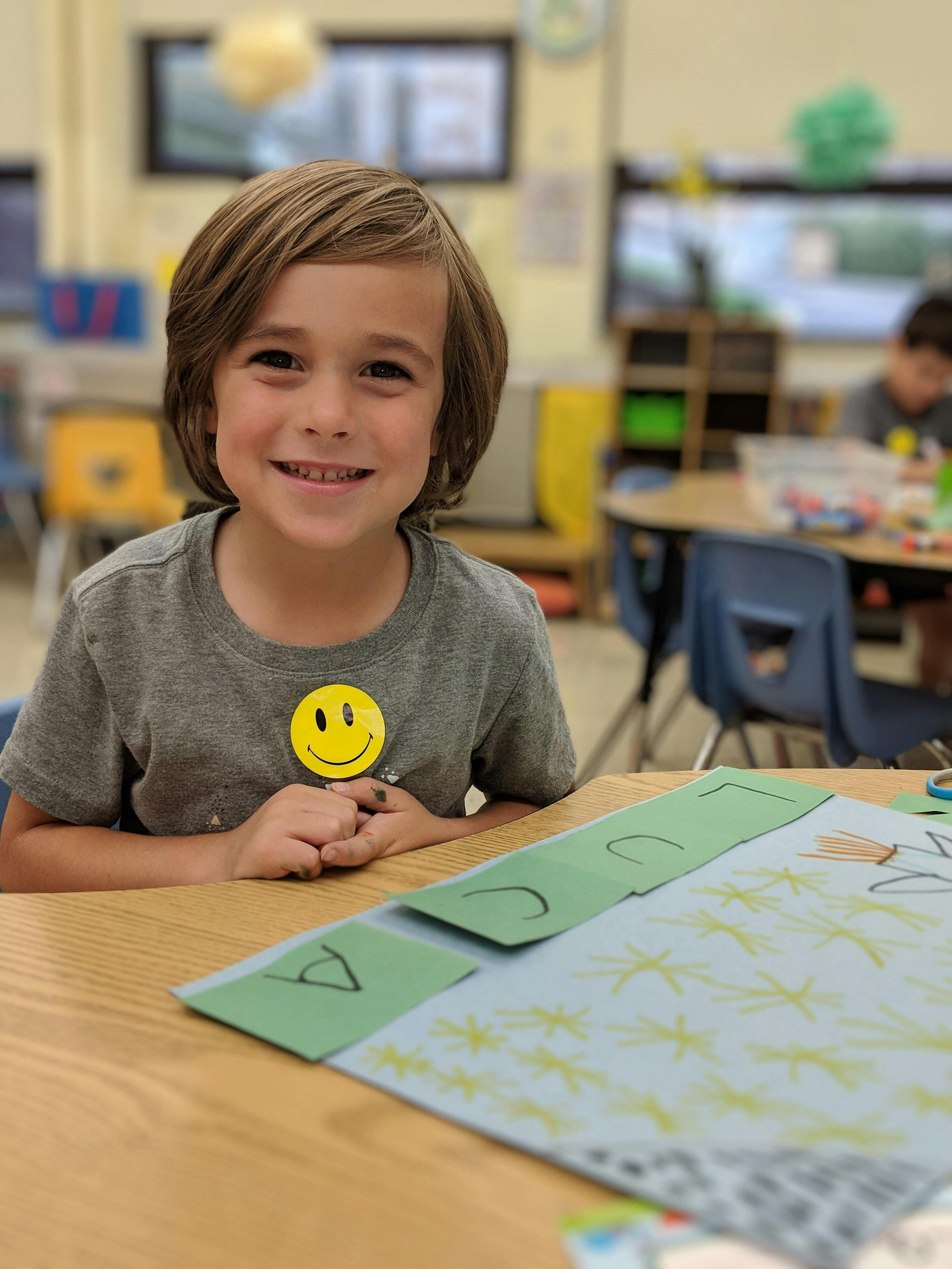 Child smiling at a desk with paper crafts in a classroom
