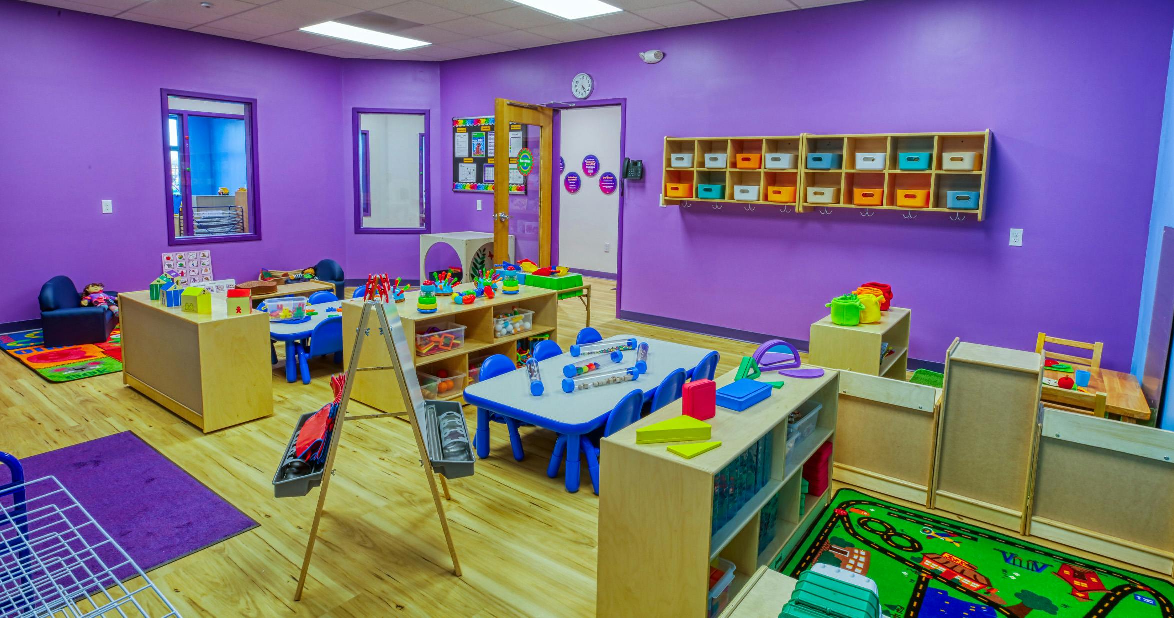 Colorful classroom with small tables and chairs, shelves with toys, and play mats