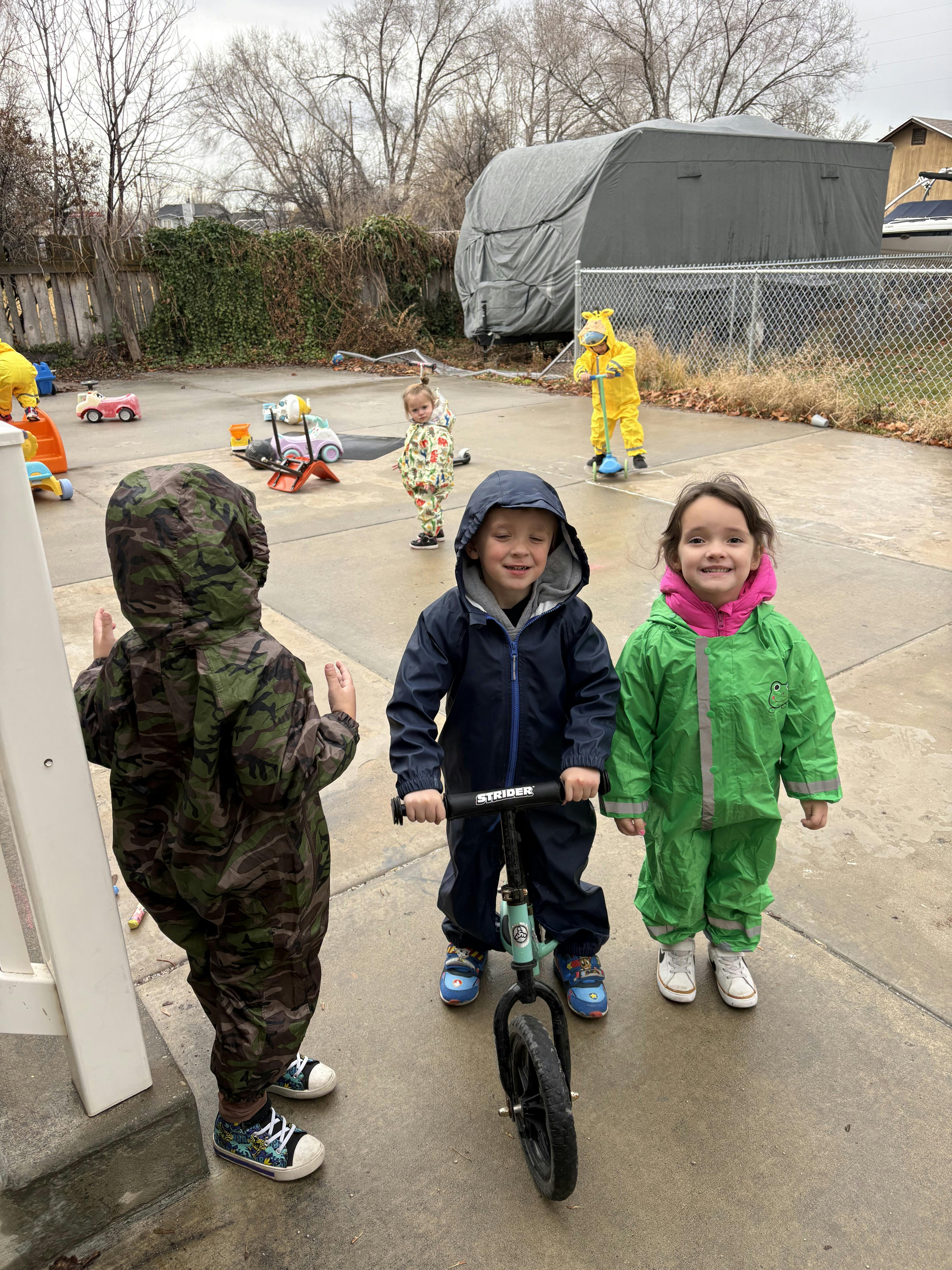 Children in rain gear playing outdoors, one on a balance bike