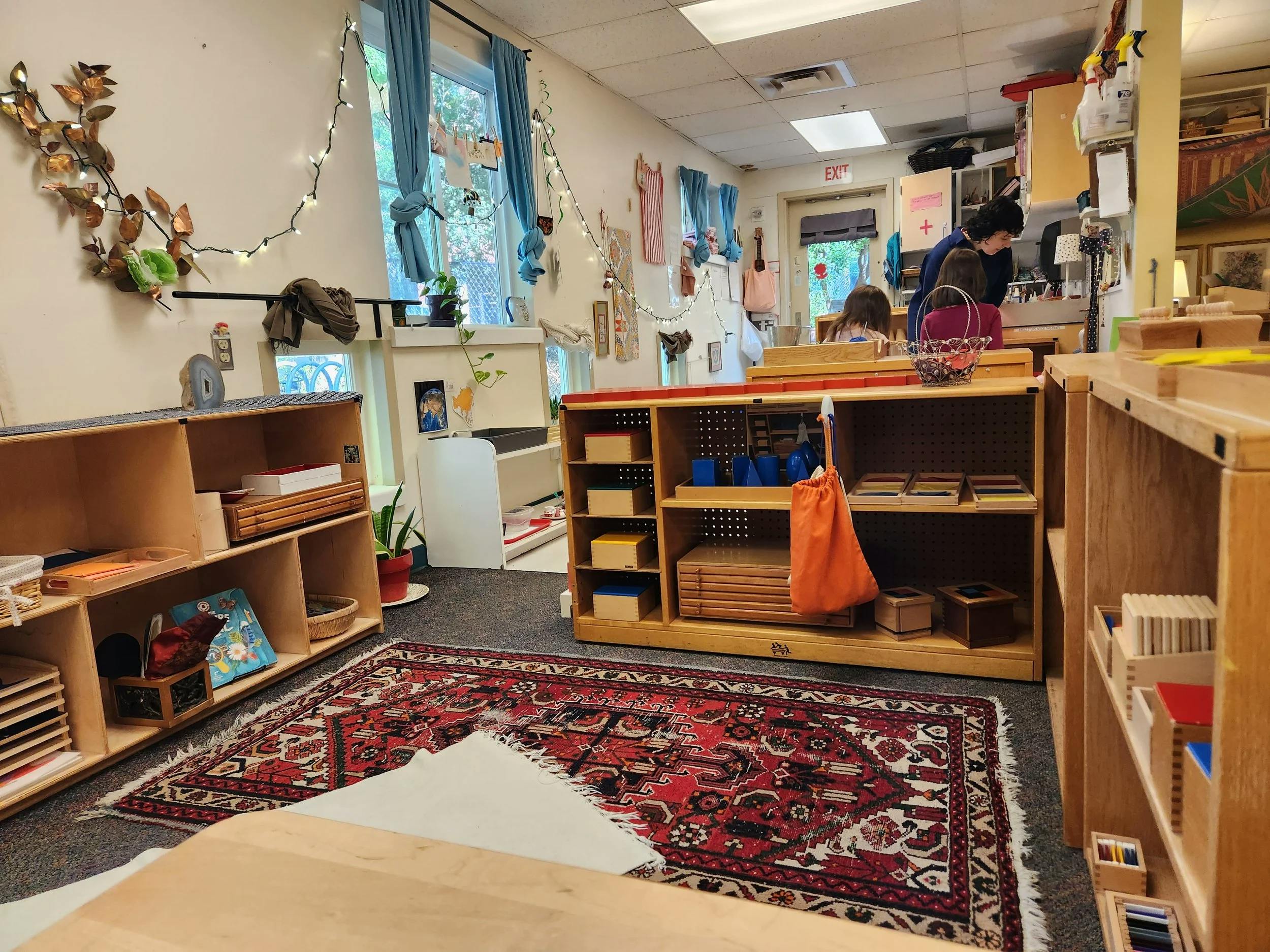 Classroom with shelves, a person and child, and a red patterned rug on the floor