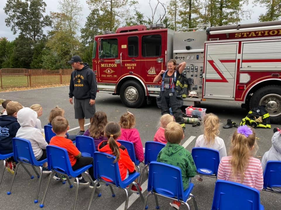Person in firefighter gear talking to children seated in front of a fire truck