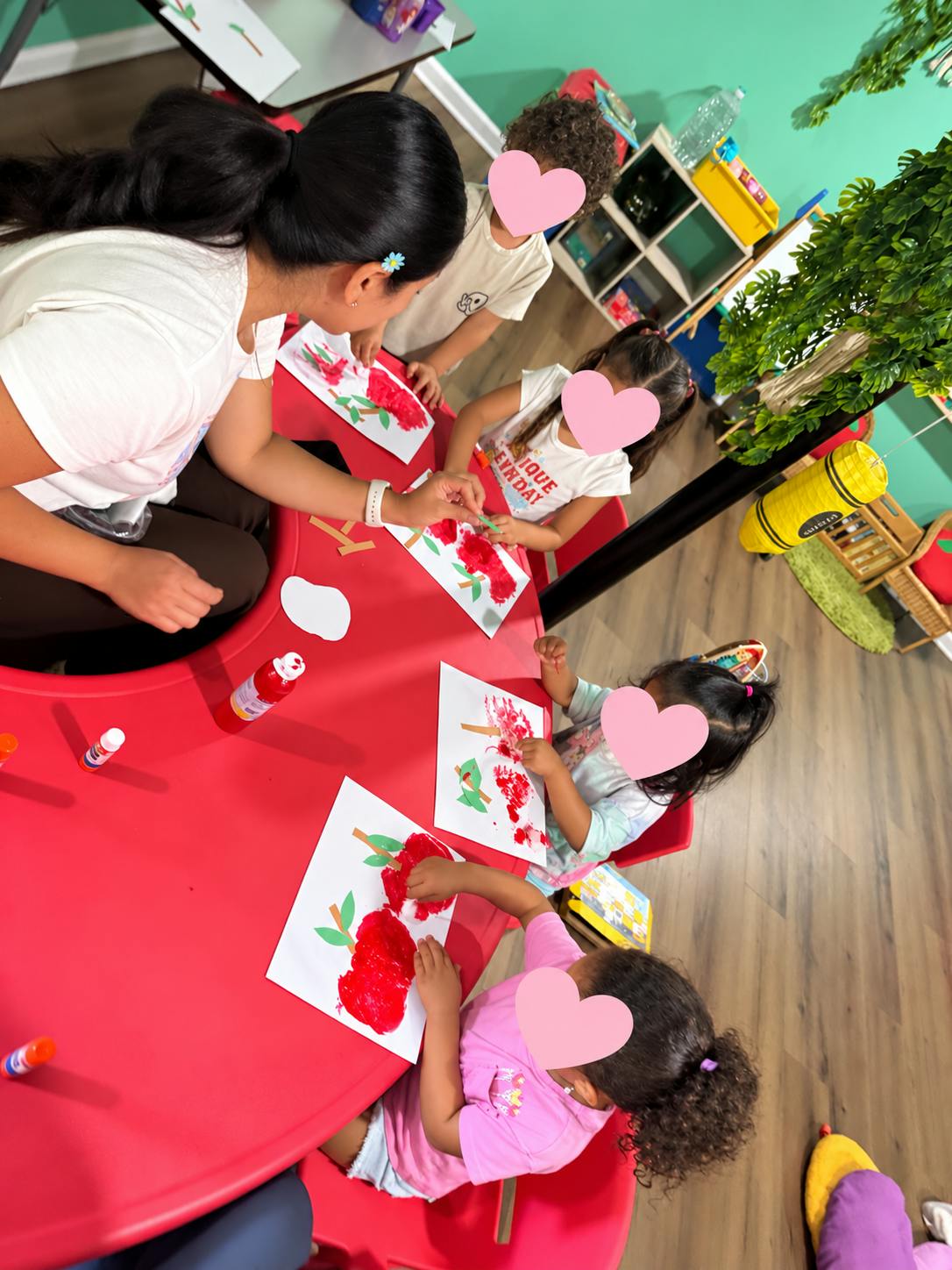 Adult assisting children with an apple craft at a red table