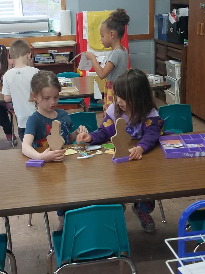 Two children at a table with craft cutouts, another child standing in background