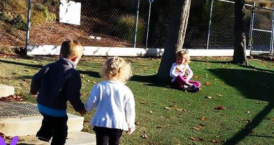Two children holding hands walking toward a child sitting under a tree