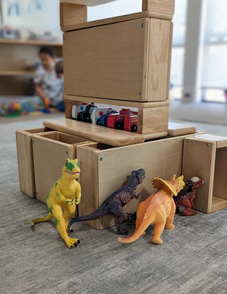 Dinosaur toys arranged in front of wooden building blocks with small toy vehicles