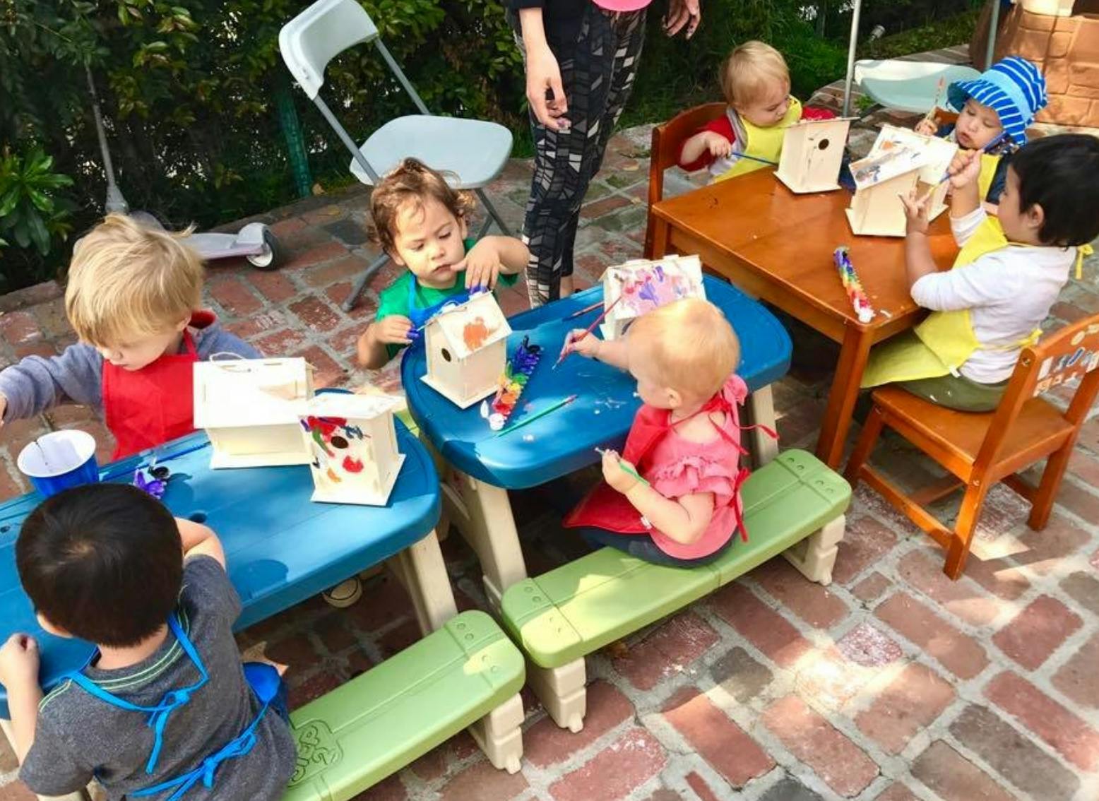 Children sitting at tables and painting birdhouses outdoors